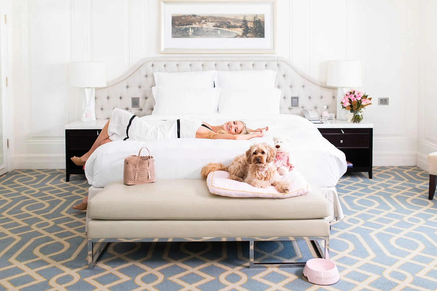A woman lying on a white bed in a bright hotel room with two dogs. One dog is sitting on a cushioned bed at the foot of the larger bed, and the woman is smiling and resting on her side. The room has white walls, a painting above the bed, bedside tables with lamps, pink flowers, a telephone, and a pink pet bowl on the floor.