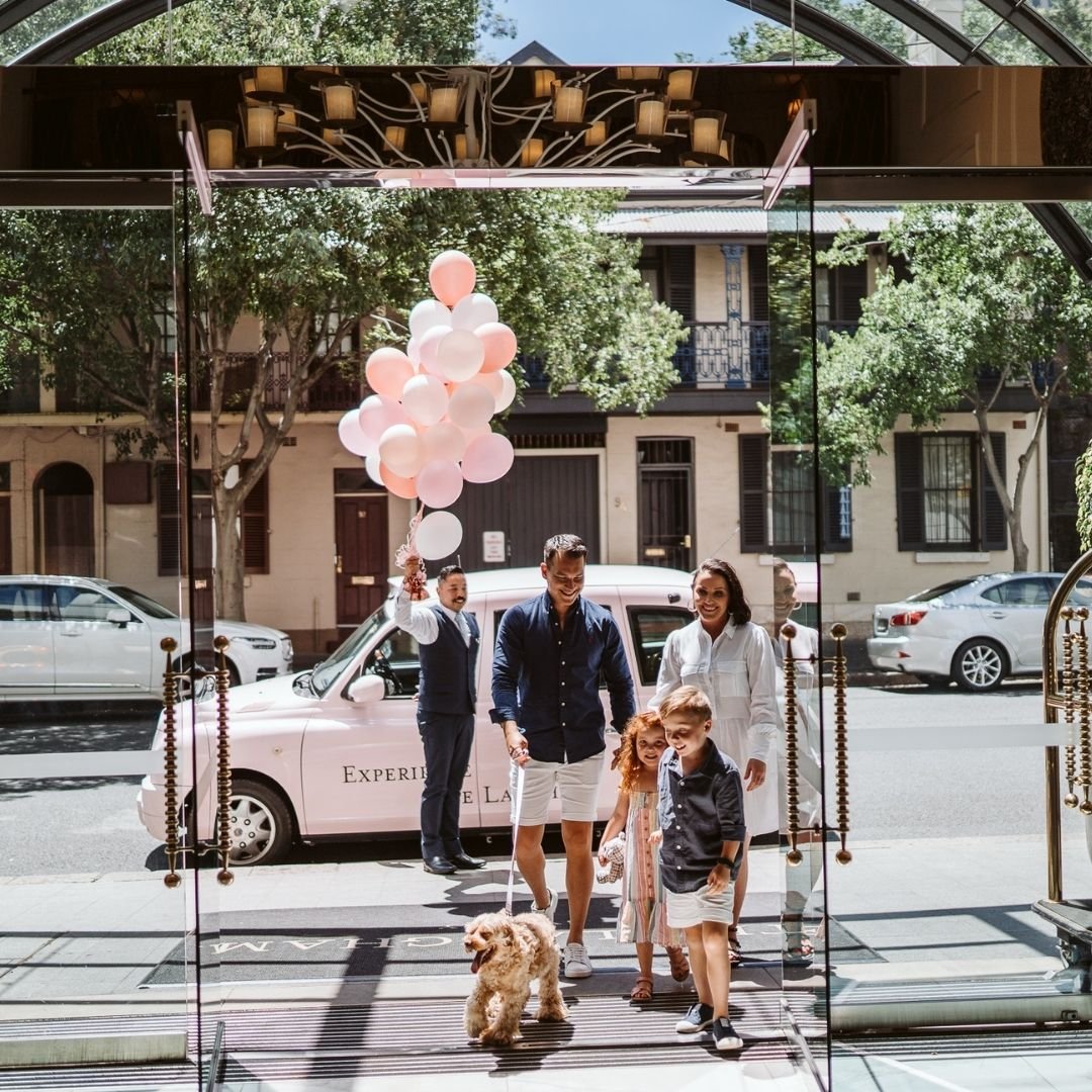 People walking out of a hotel entrance with balloons, a dog, and friendly smiles on a sunny day.