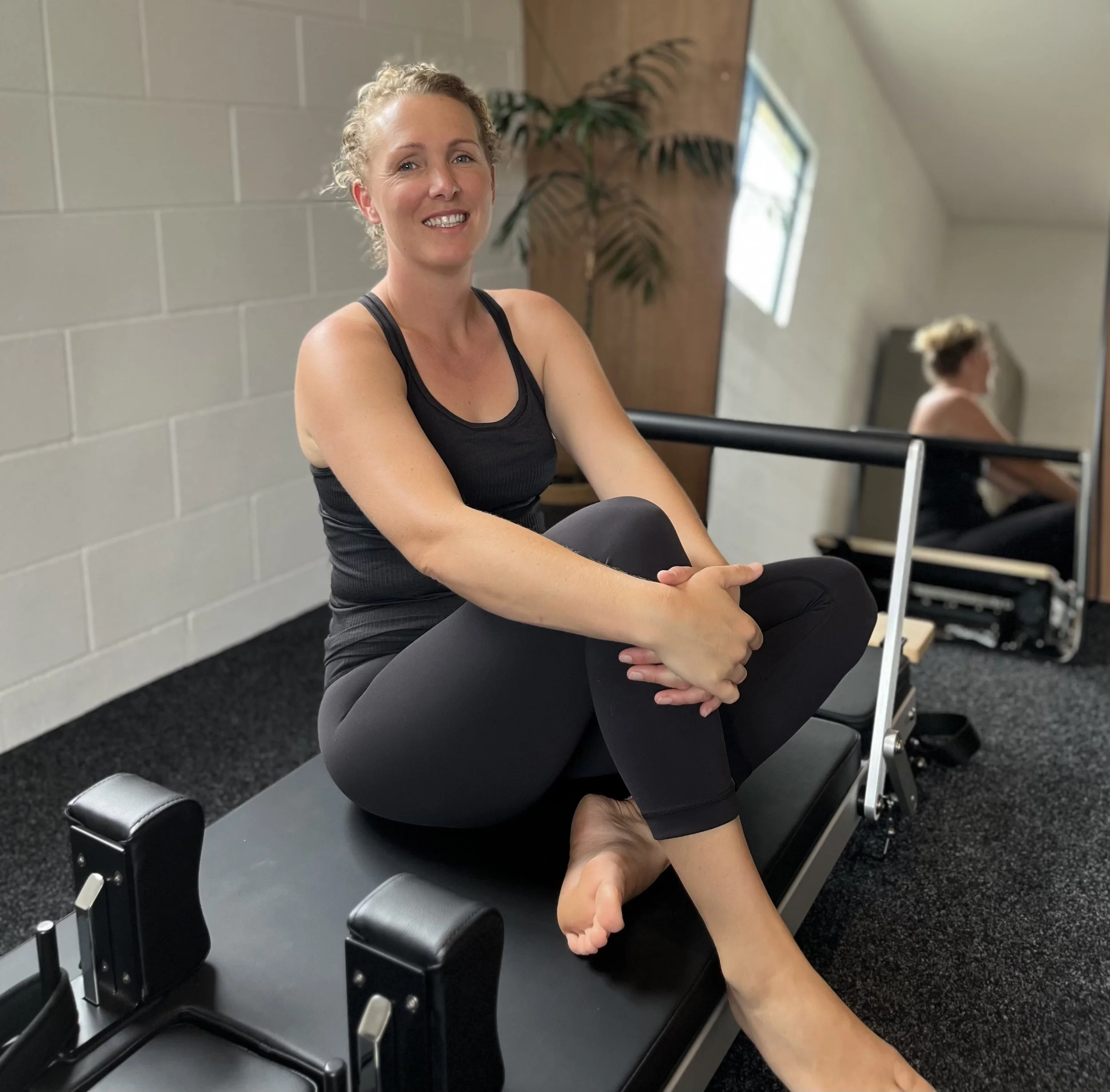 A woman sitting cross-legged on Pilates reformer equipment in a fitness studio, smiling at the camera. She is wearing a black tank top and black leggings, with mirror and window reflections in the background.