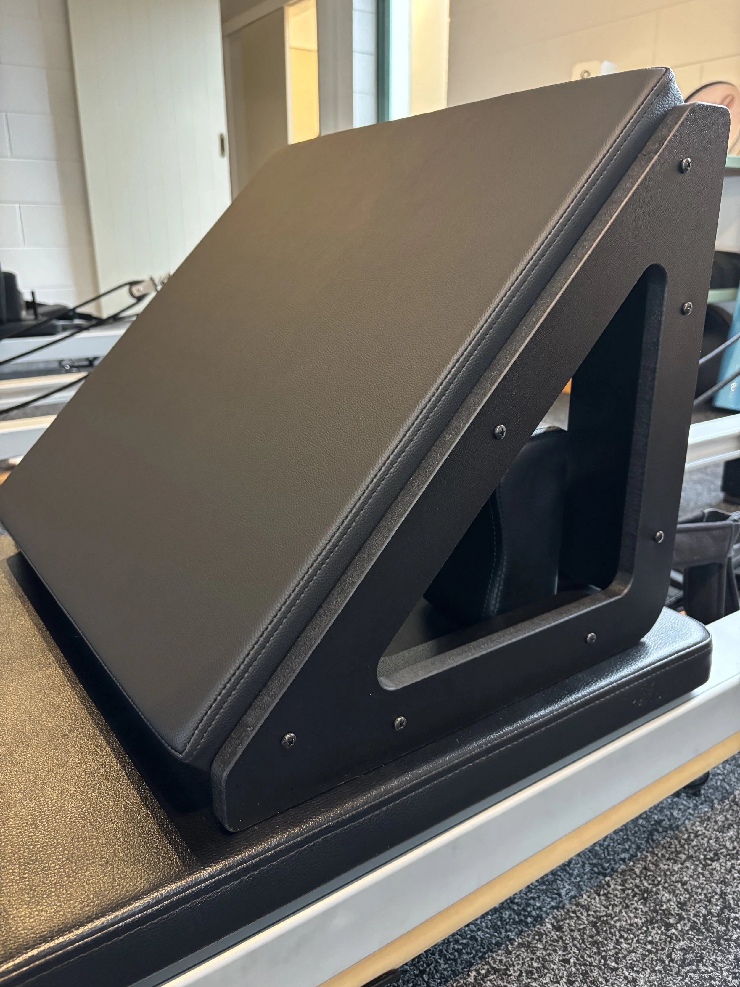 A black back board of a reformer or physical therapy treatment table with an adjustable headrest positioned at an incline, located in a room with beige brick walls and a window.
