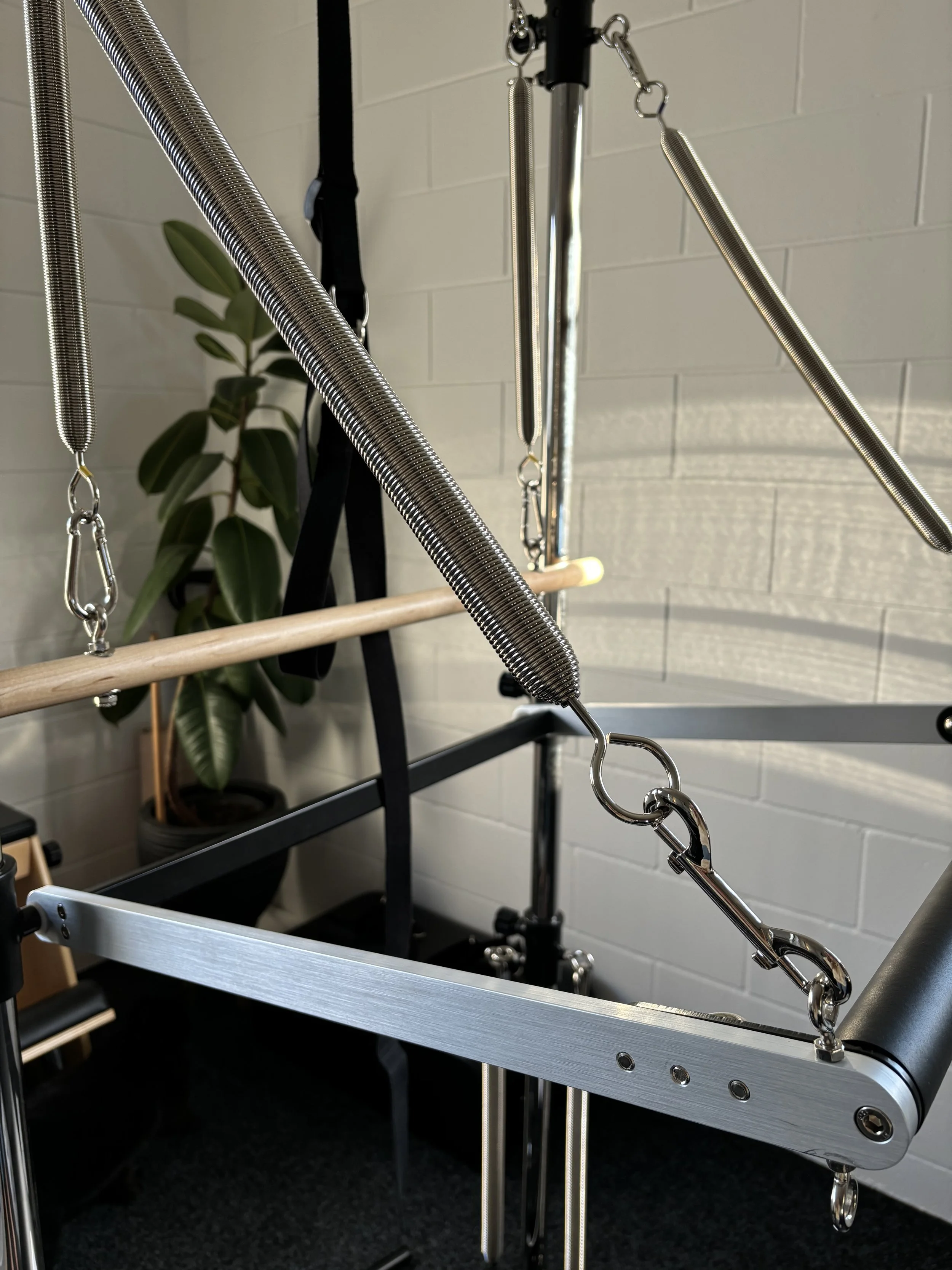 A Pilates Cadillac reformer machine with metal springs and attachment hooks in a fitness studio, with a potted plant in the background.