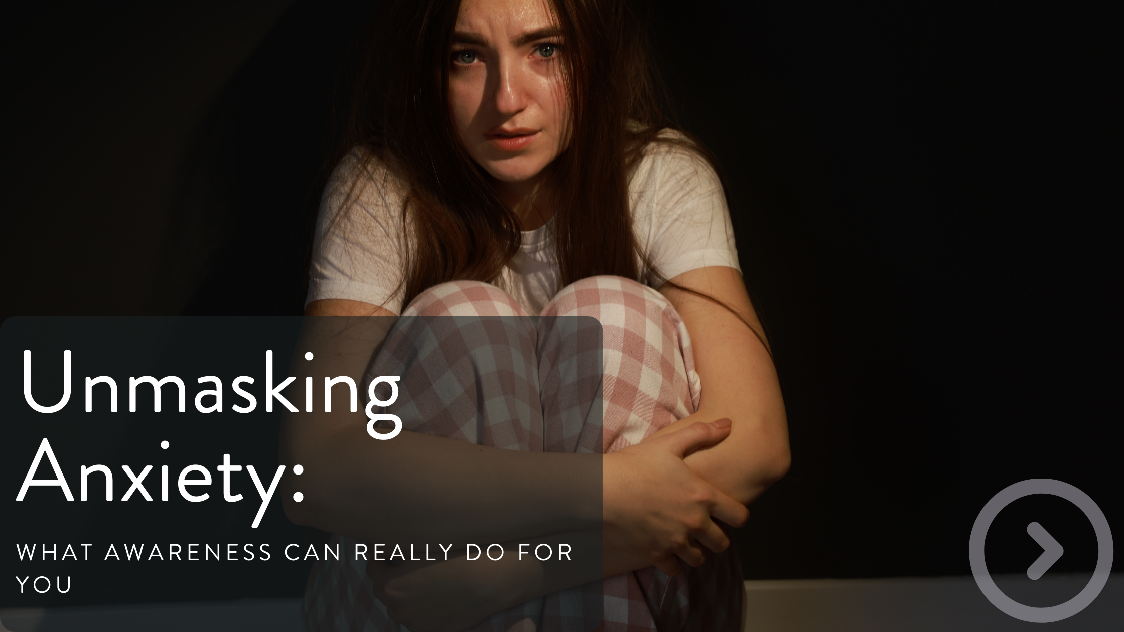 A woman with long red hair sitting on the floor, hugging her knees, and looking distressed against a dark background.