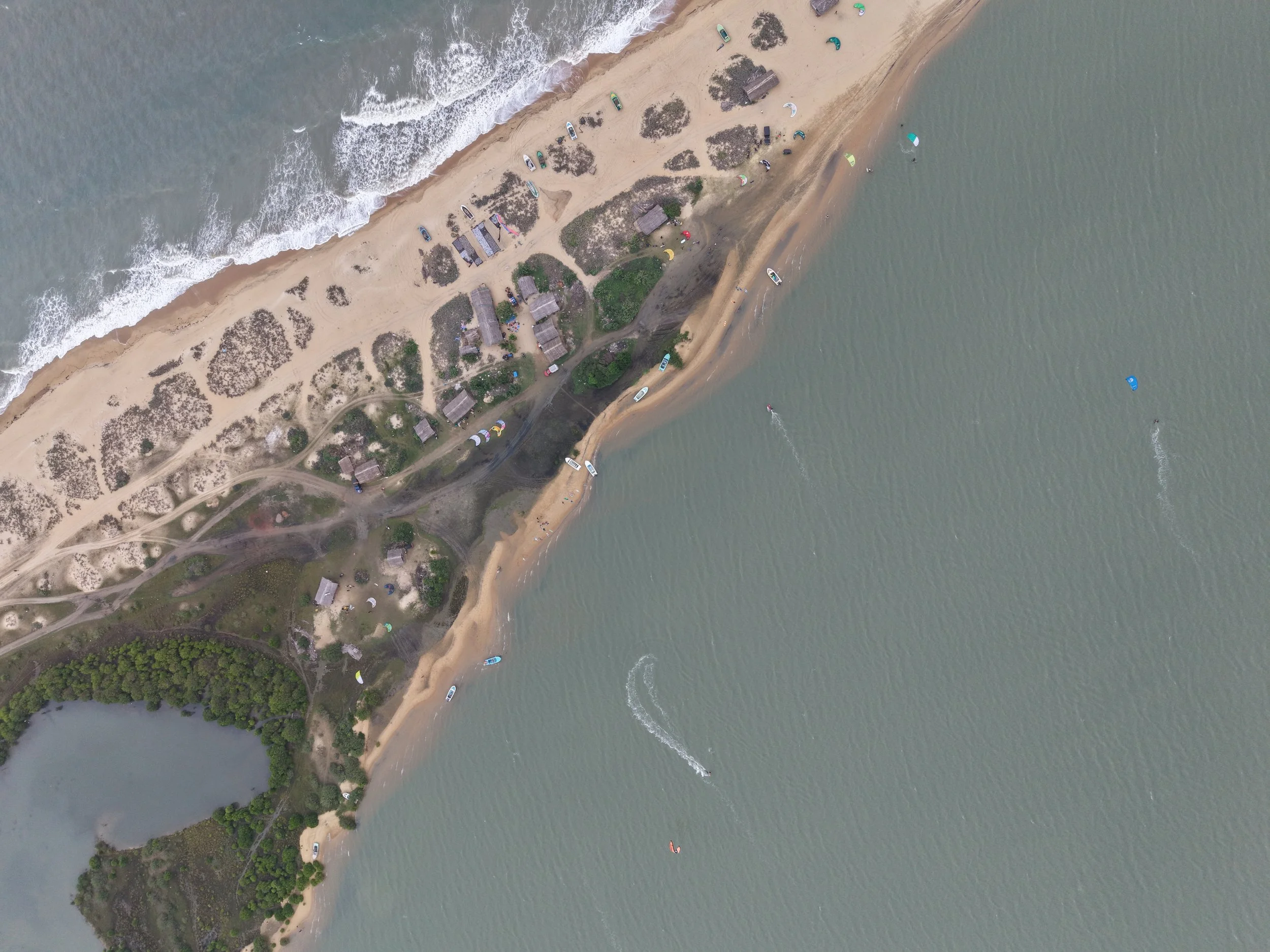 Aerial view of a coastal area with a sandy beach, small buildings, and boats on the water, some with windsurfers.