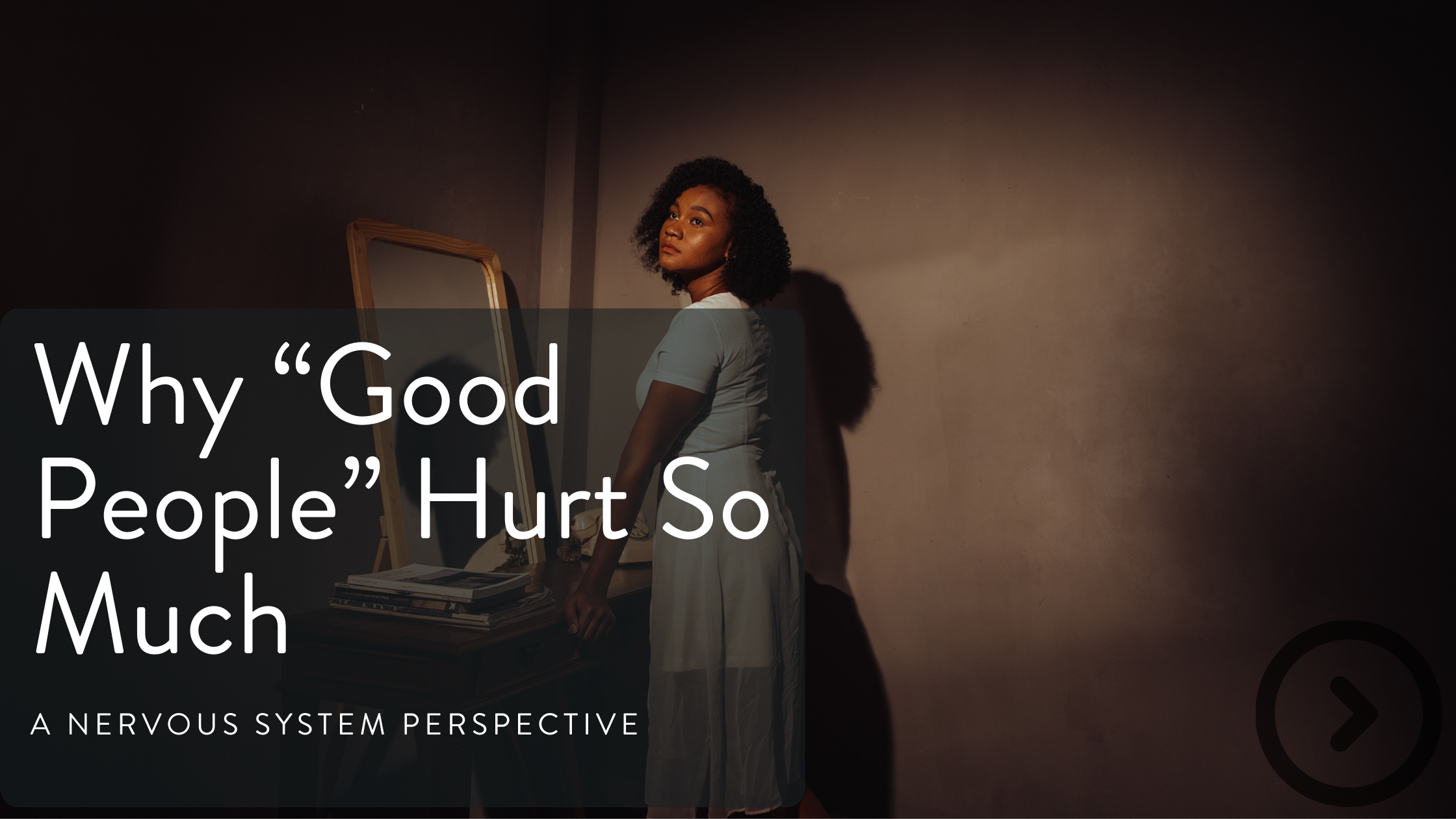 A woman with curly hair standing in a dimly lit room, looking pensively at a mirror, with a stack of books on a table beside her and a shadow cast on the wall.