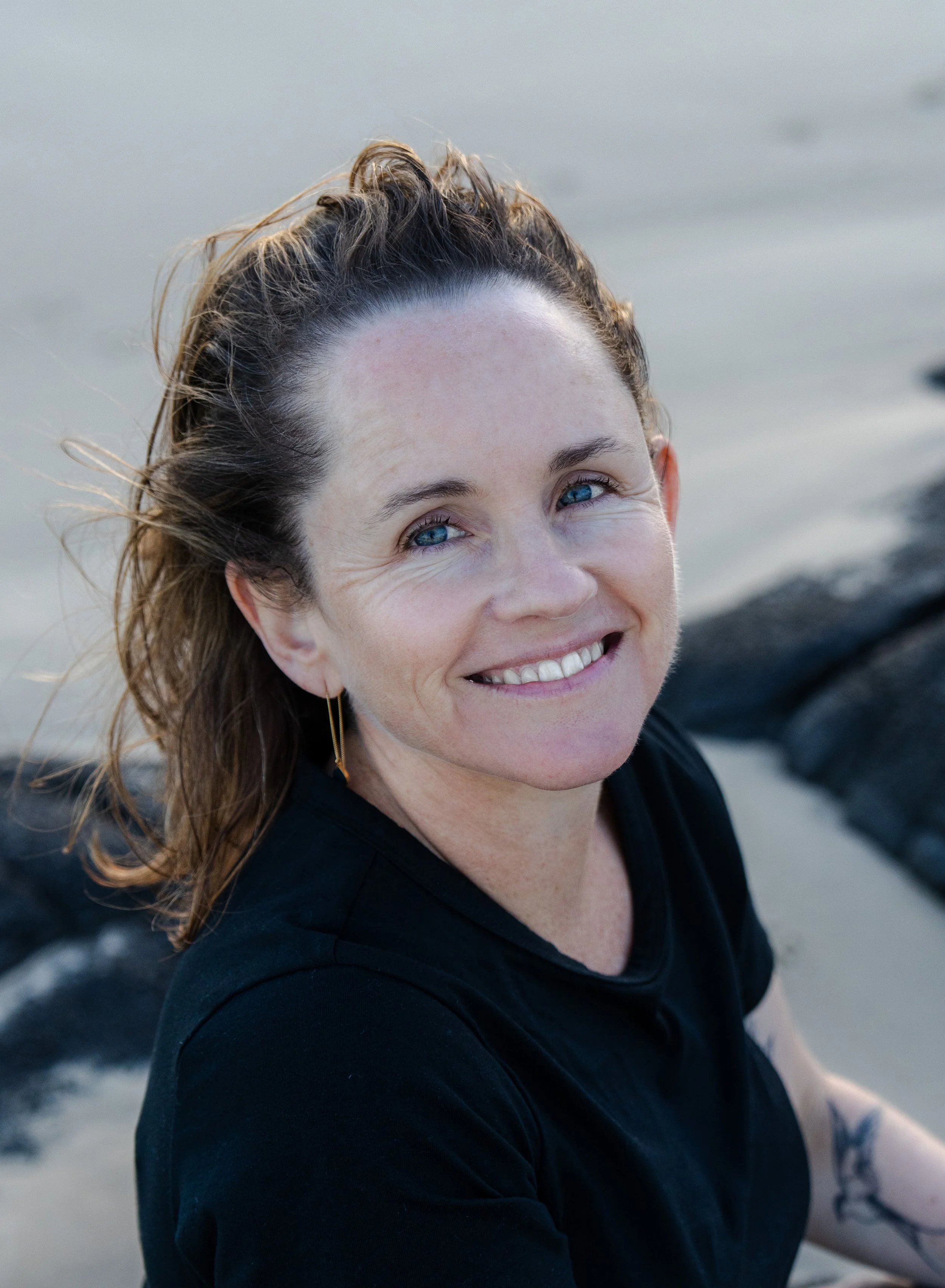 Fiona MacLeod Close-up of a smiling woman with blue eyes and brown hair, outdoors near water and rocks, wearing a black shirt.