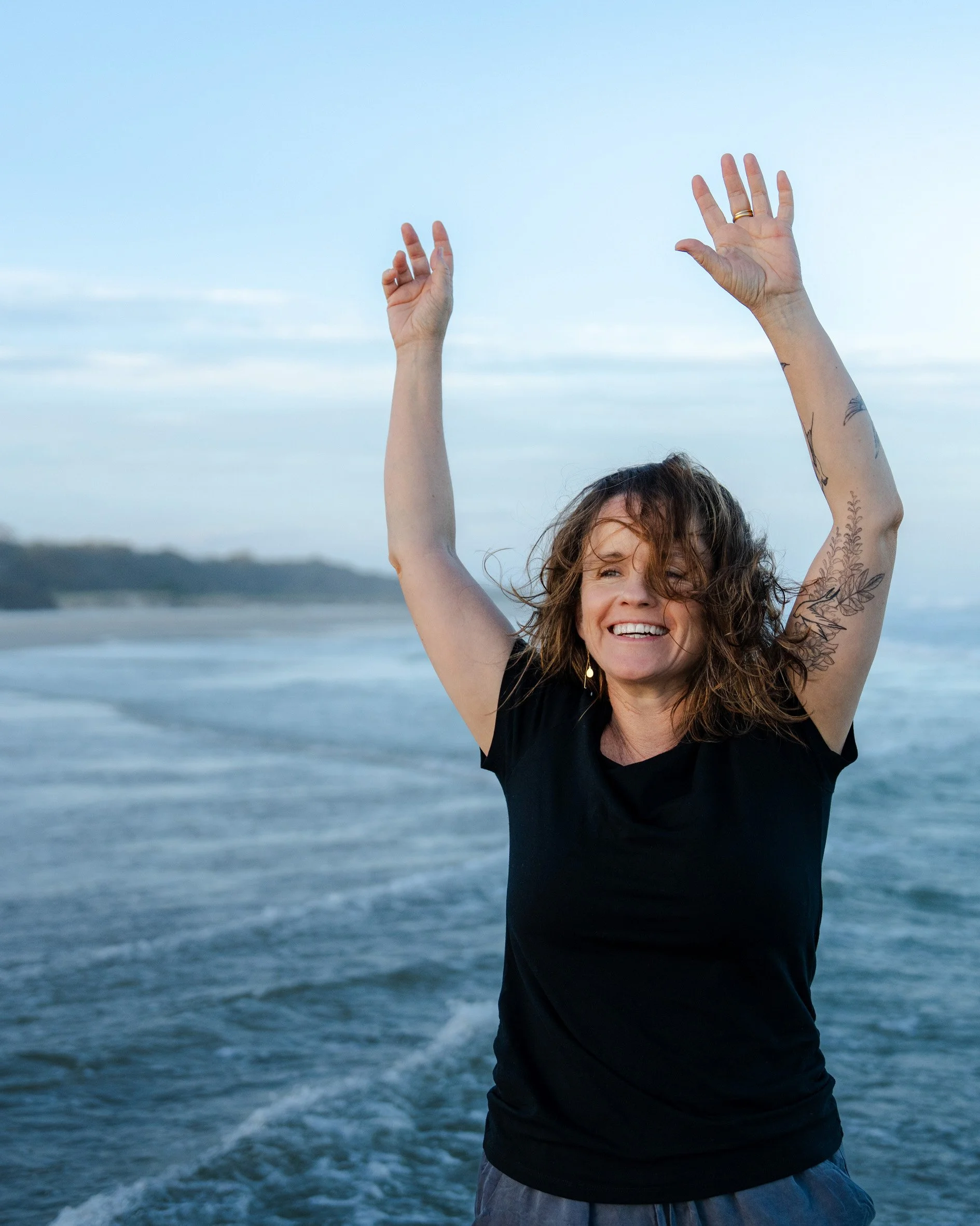 Fiona on the beach at sunset, smiling, happy, arms reaching up. Therapeutic movement in nature