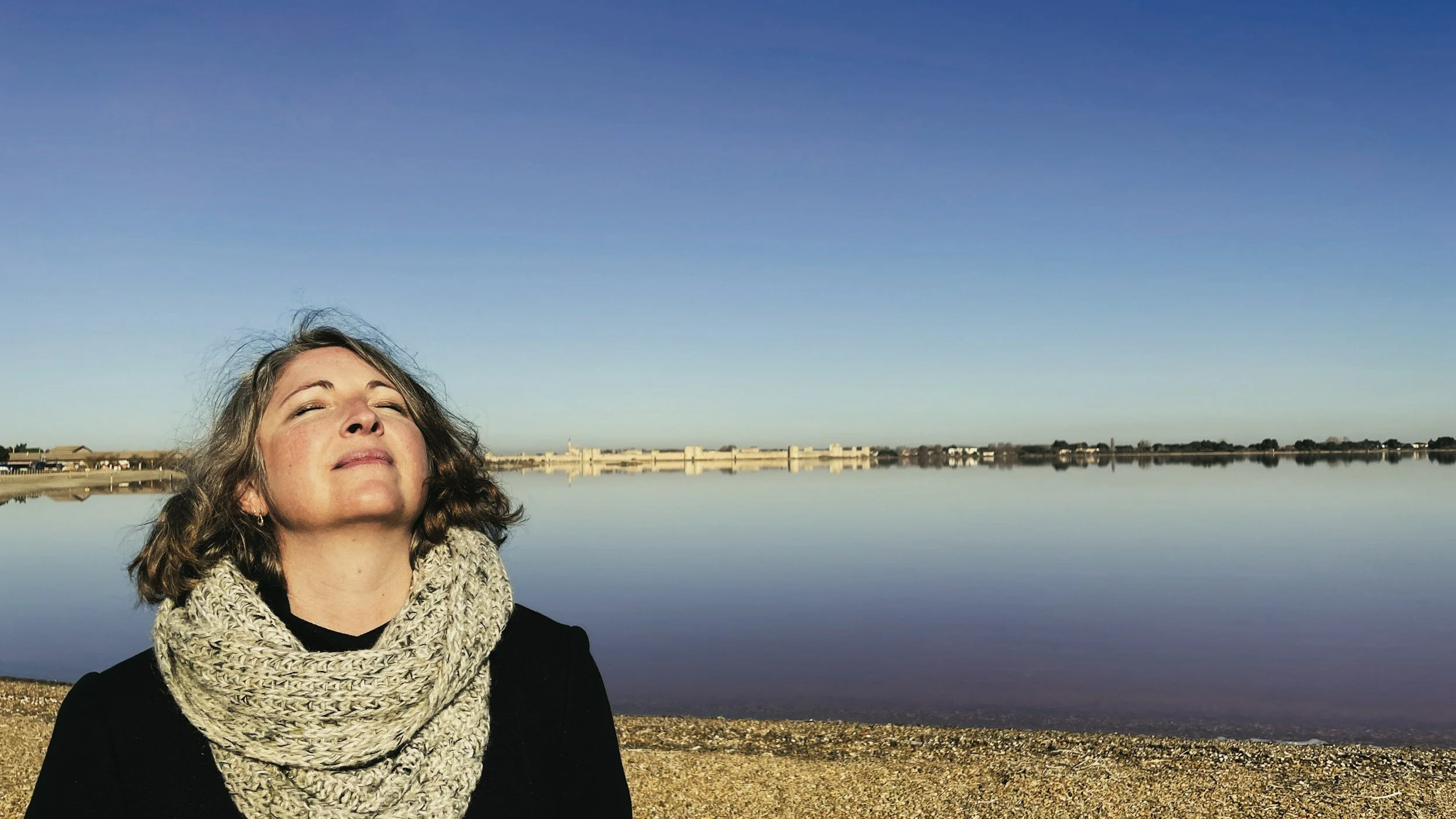 Woman with curly hair and a beige knitted scarf outdoors with a large body of water and a distant shoreline under a clear blue sky.