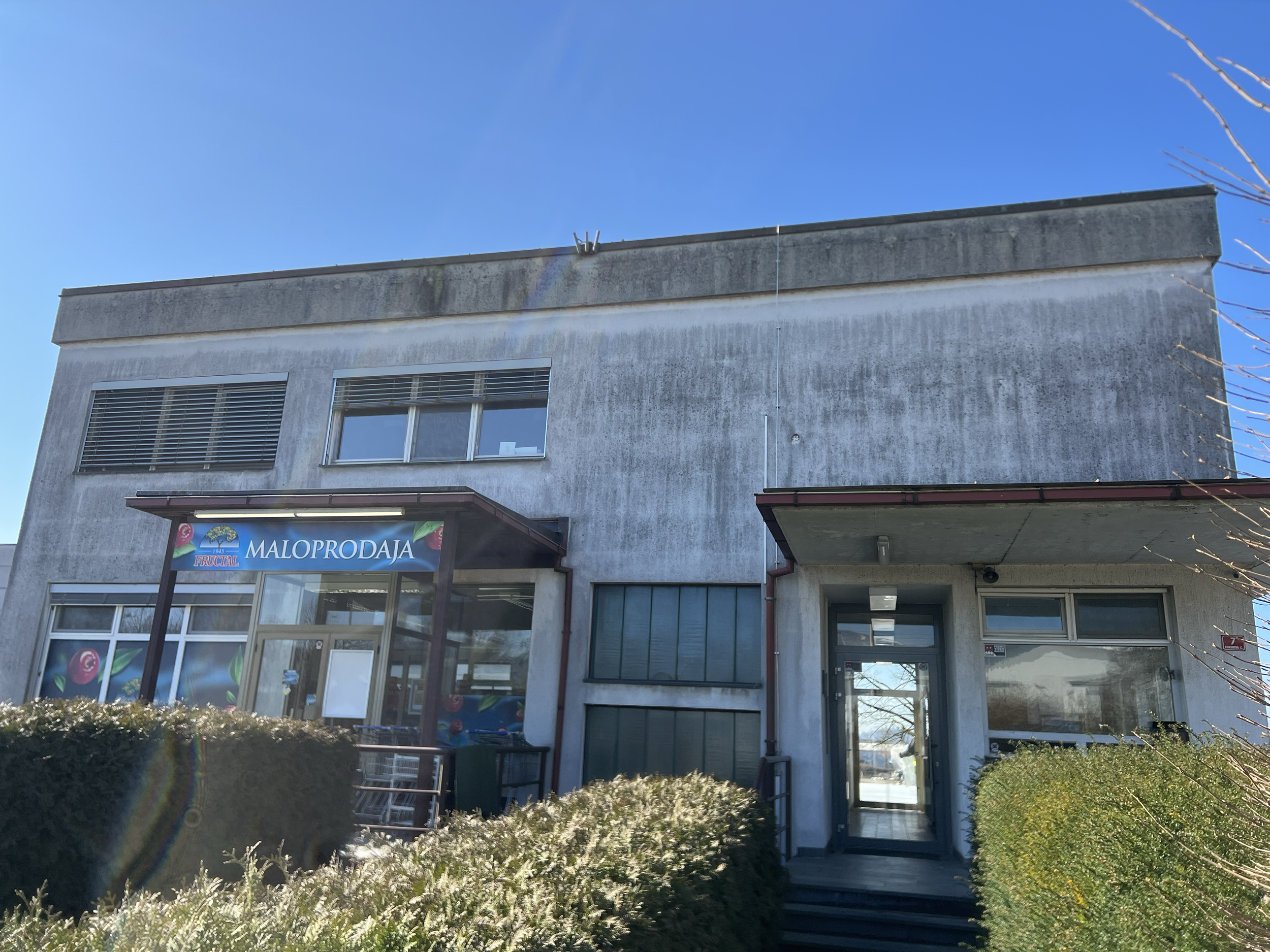 Front view of a commercial building with signs, windows, and entrance, with bushes and plants in front, under a clear blue sky.
