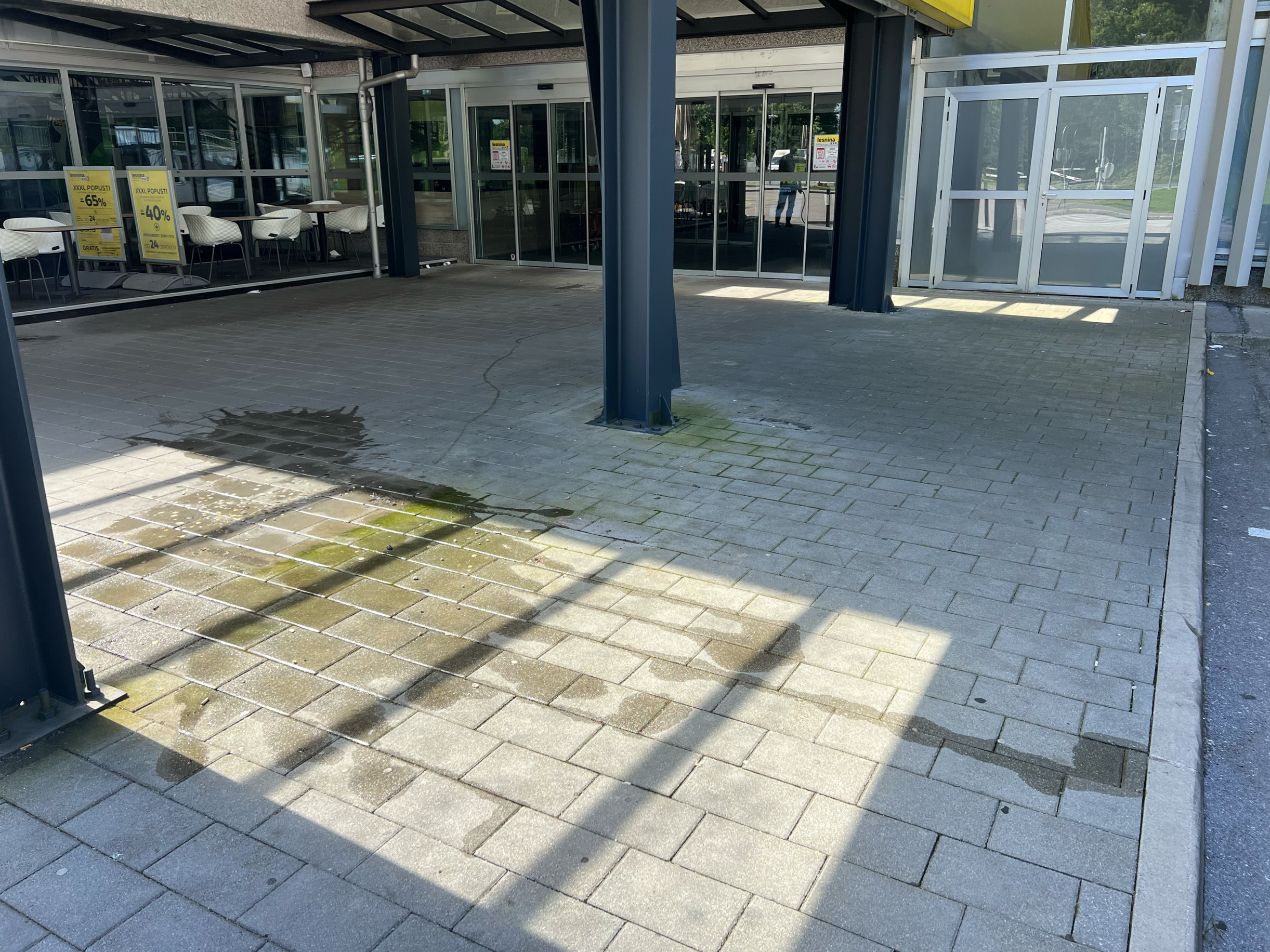 Exterior view of a commercial building with glass doors and windows. There are several yellow sale signs in the windows advertising discounts. The area has paved ground with some water puddles and shadows from nearby objects.