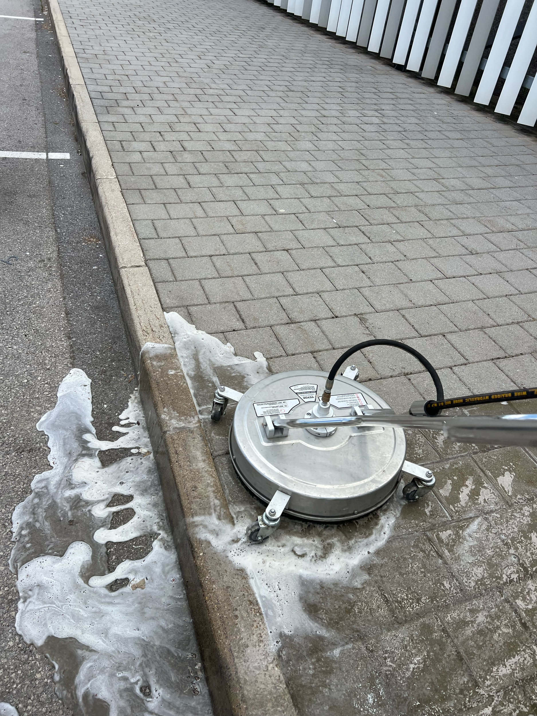 A sidewalk pressure washer cleaning the curb and pavement near a building with metallic slats.