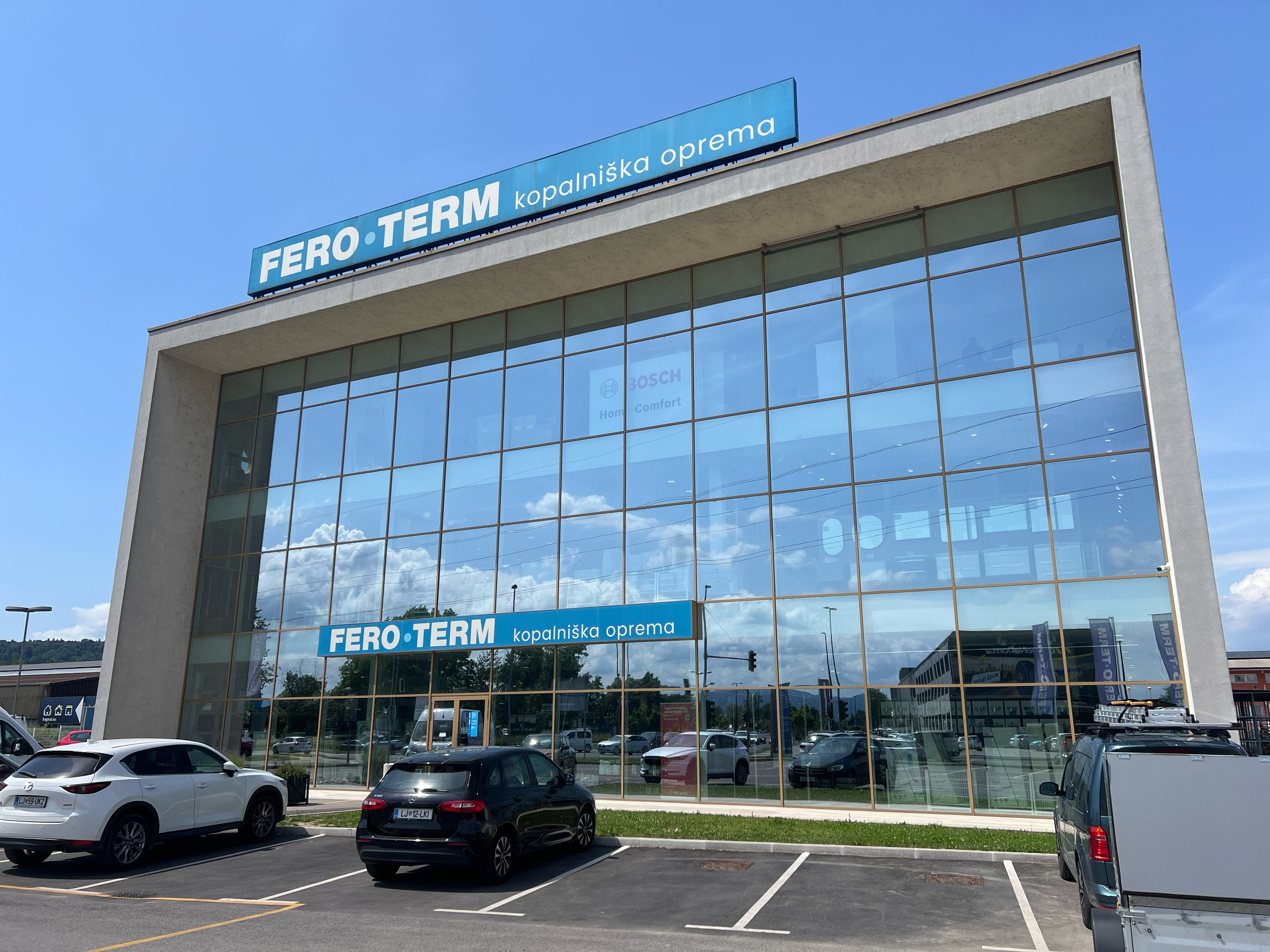 Modern multi-story building with large glass windows and a blue sign on top that reads "FERO TERM kopalniška oprema". There is a parking lot with several cars in front of the building under a clear sky with some clouds.