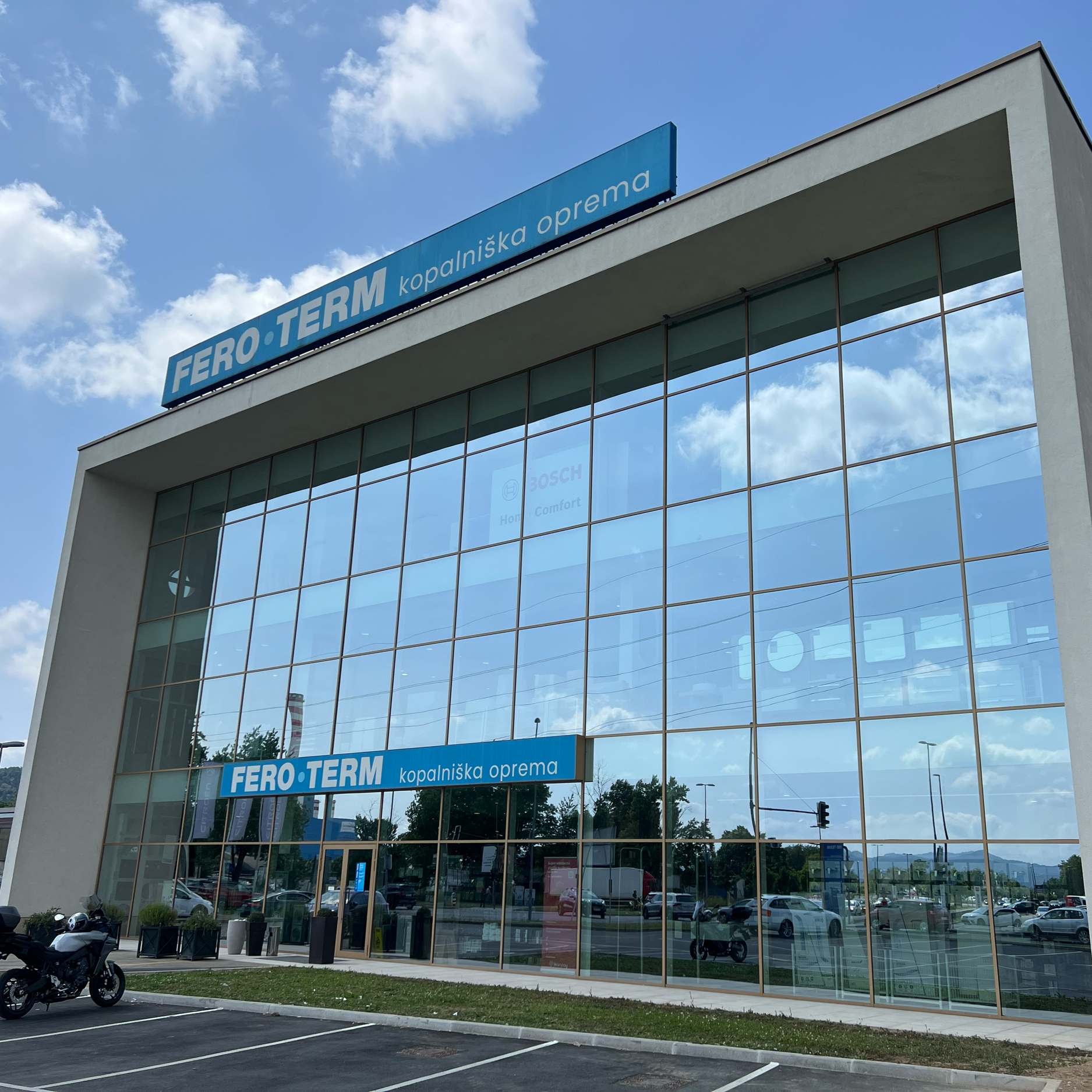 Modern building with large glass windows and blue signs that read "FERO TERM kopalniška oprema" against a partly cloudy sky. Parking lot with cars and motorcycles in front of the building.