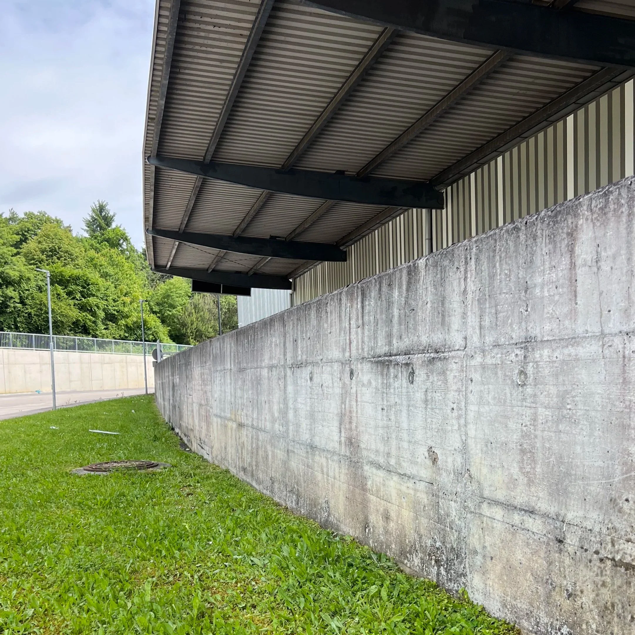 Concrete wall with a metal awning overhead, grassy area at the base, and a background of trees and streetlights.