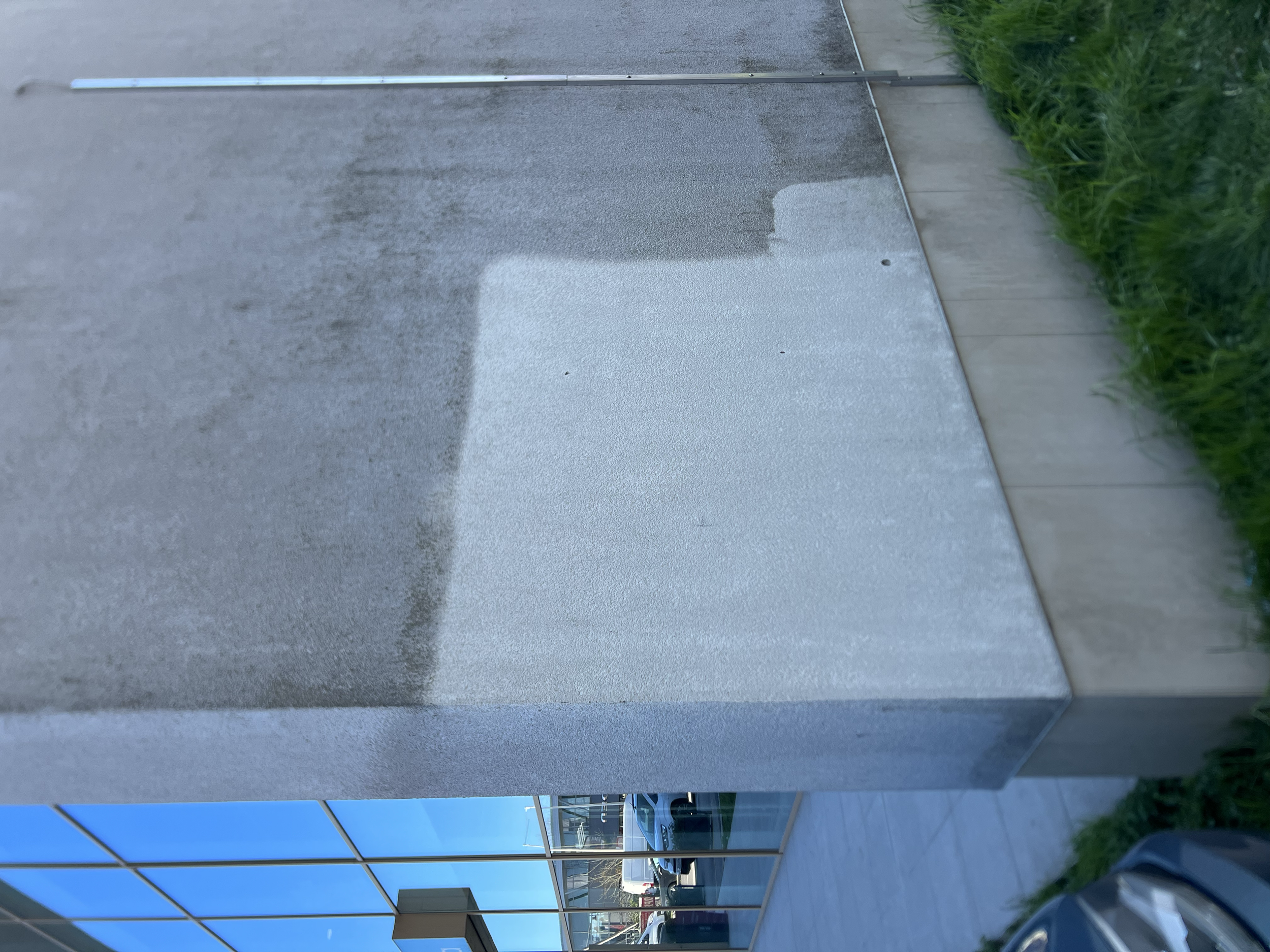 Close-up of a concrete sidewalk with darker and lighter sections, bordered by grass and a glass building in the background.
