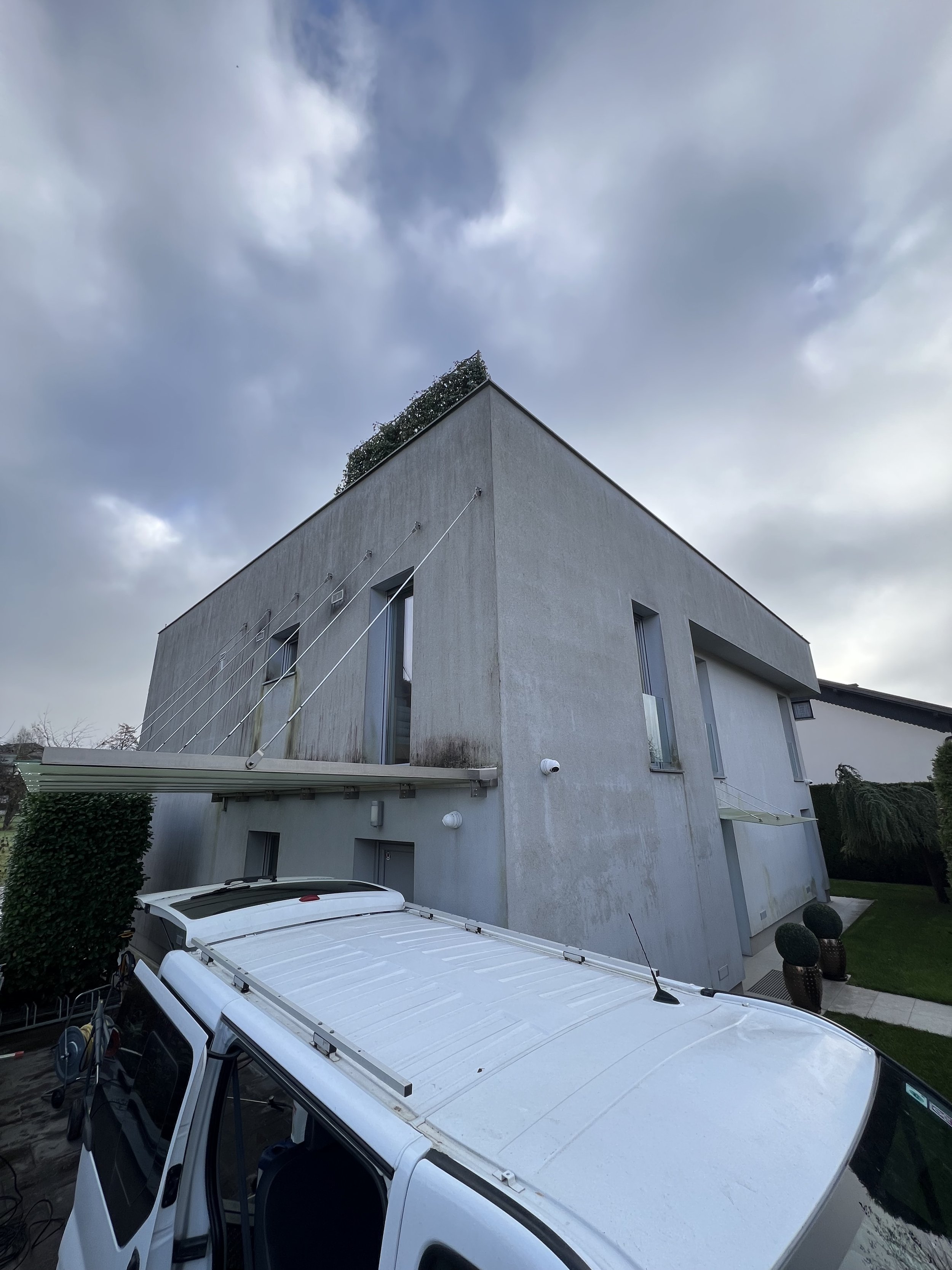 Modern gray house with rectangular windows, a white vehicle in front, and a cloudy sky overhead.