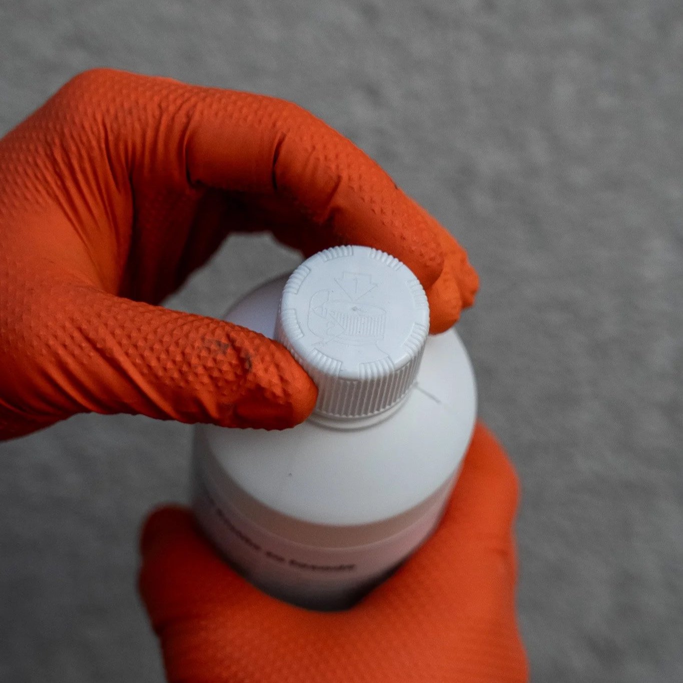 A person wearing orange gloves holding a white bottle with a white screw top cap. The background is a gray surface.