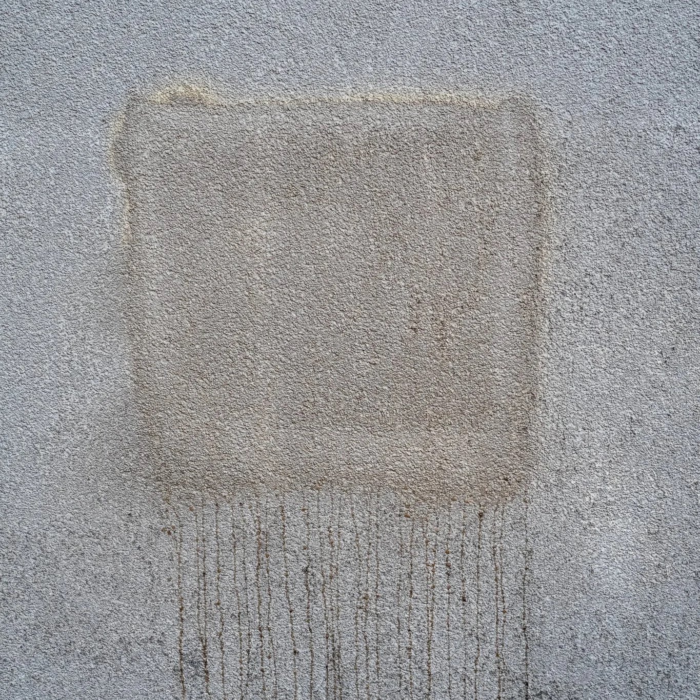 Close-up of a textured concrete wall with faint graffiti. The wall has a rough surface with some streaks of dirt or weathering.
