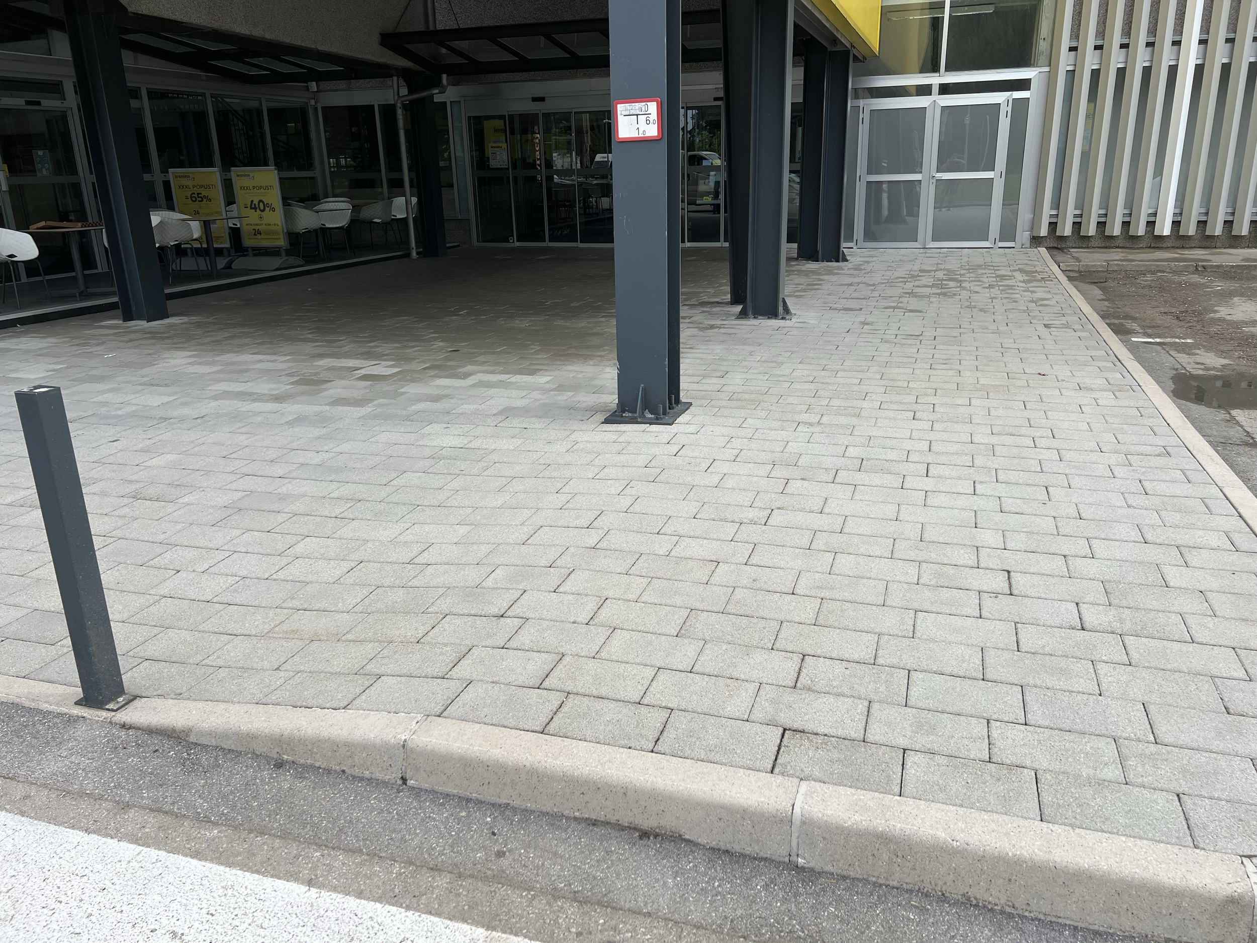 Exterior view of a modern commercial building with glass doors and windows, a paved sidewalk, and signs advertising discounts. There are some plastic chairs inside visible through glass.