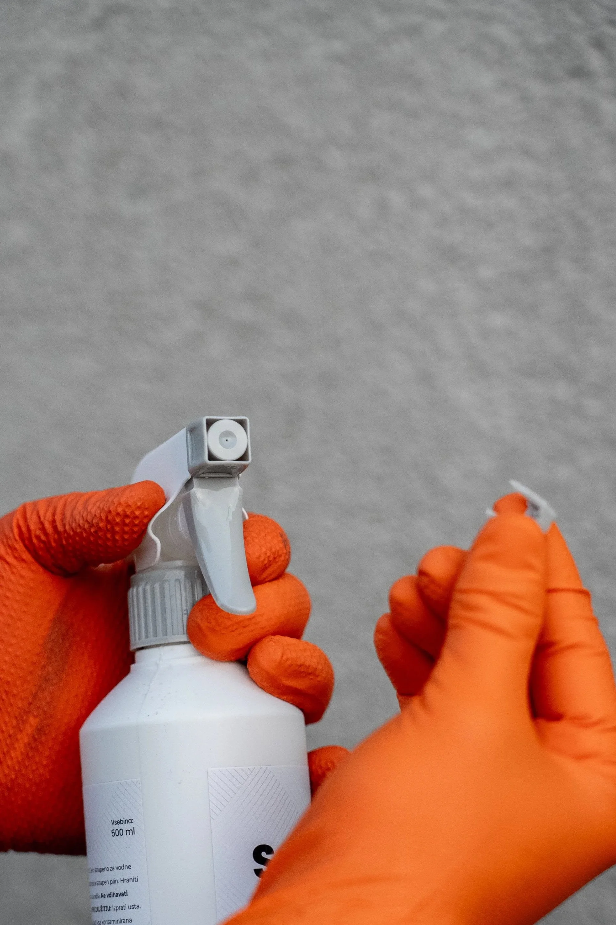 Person wearing orange gloves holding a white spray bottle, with a gray ceiling in the background.