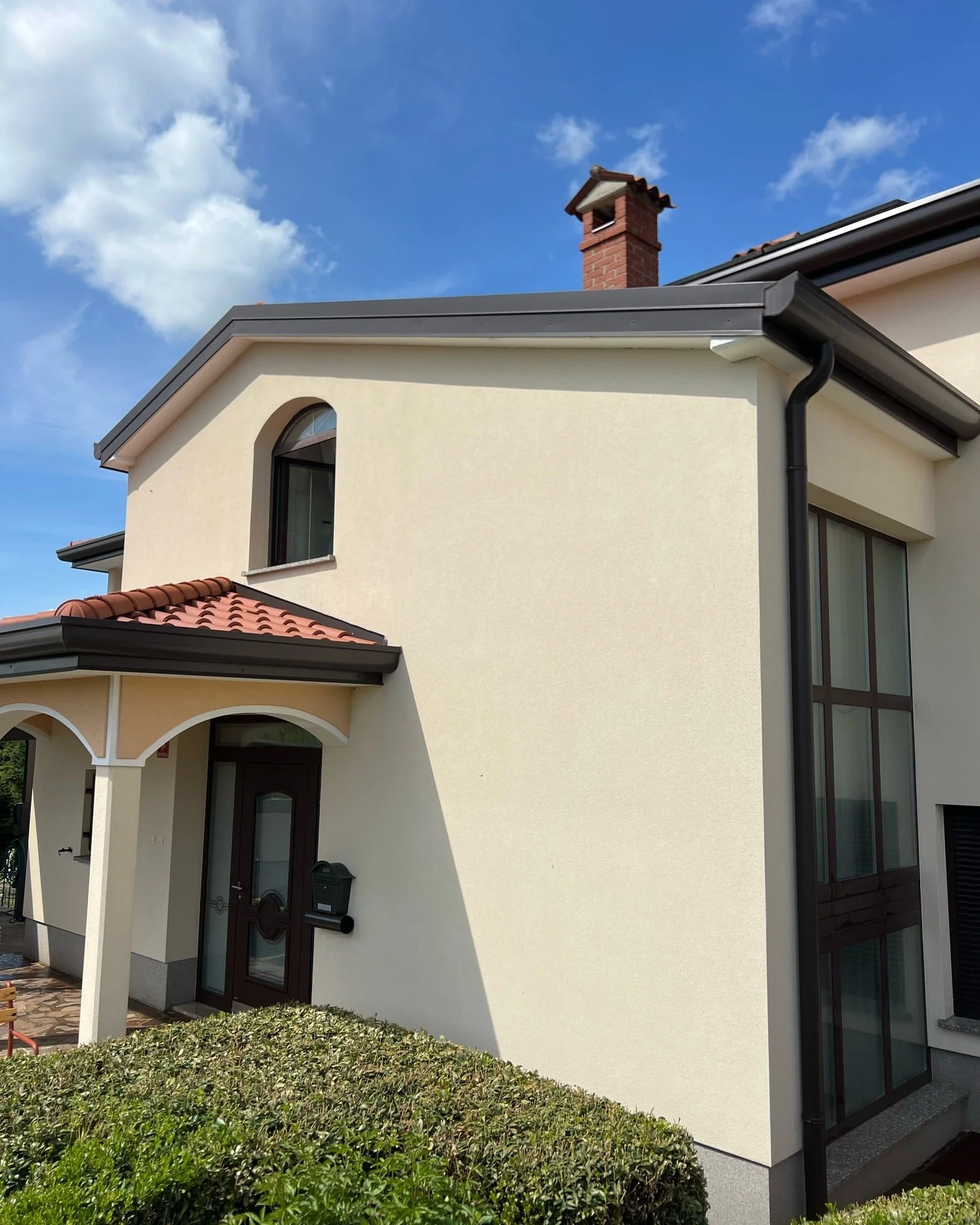 Exterior of a modern house with light-colored walls, a red tile roof, a brick chimney, and large windows, with a clear blue sky with some clouds in the background.