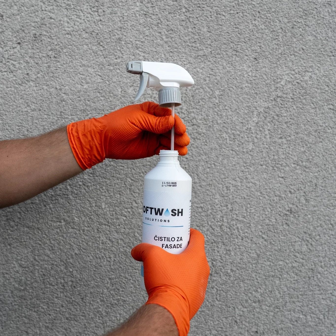 Person wearing orange gloves holding a spray bottle labeled 'SoftWash Solutions' with the text 'Cistilo za fasade' in front of a textured gray wall.