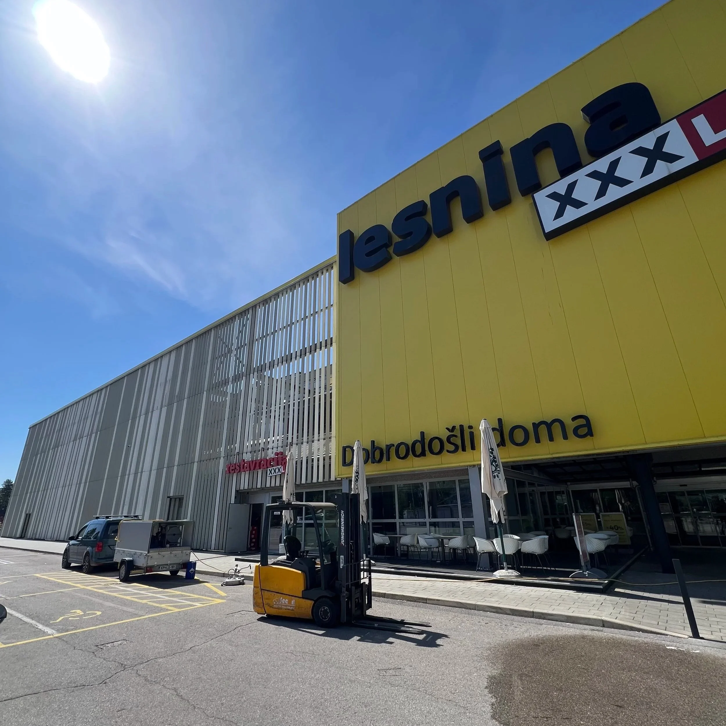 Large yellow building with a sign reading 'Lidl' and additional text in Croatian, adjacent parking lot with a white utility trailer, forklifts, and umbrellas for outdoor seating.