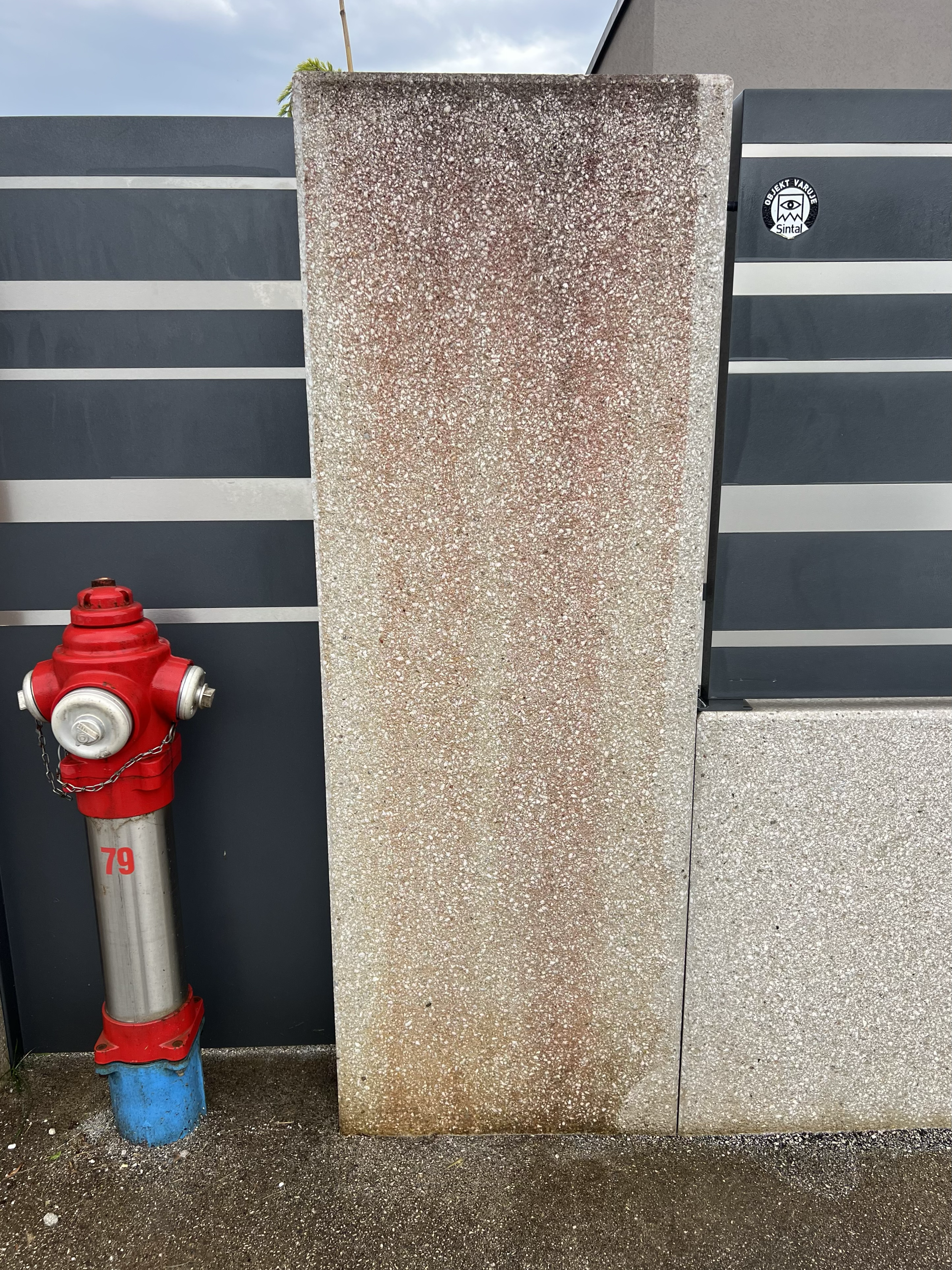 A large textured rectangular stone or concrete slab leaning against a dark gray fence, with a red fire hydrant nearby on the ground.
