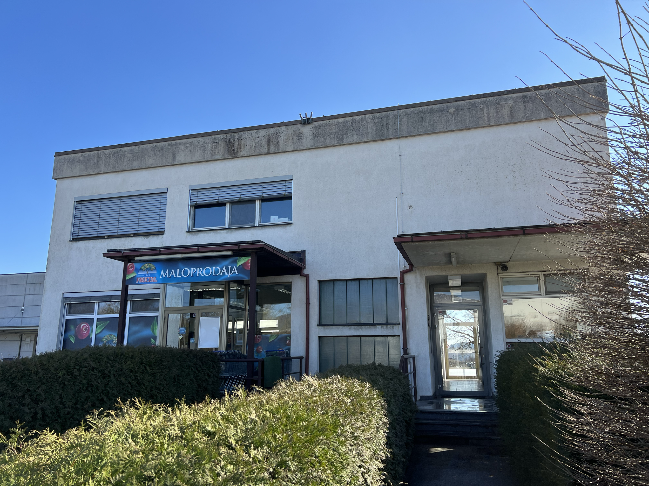 The front of a two-story commercial building with a supermarket named Maloprodaja Frucal. The building is white with two horizontal windows and a large storefront window. The sky is clear and blue, with some trees and greenery in front.