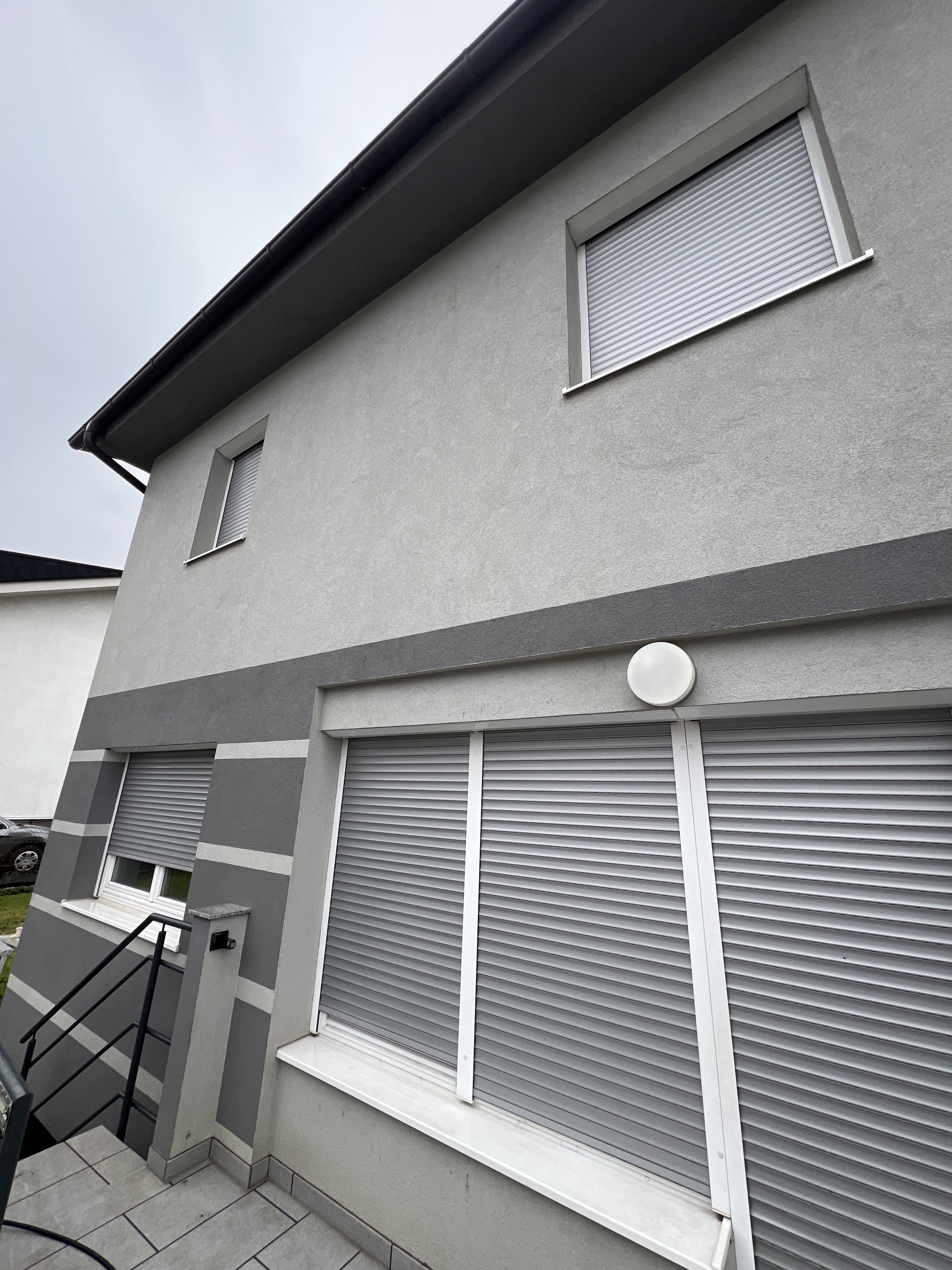 Exterior of a modern house with light gray walls, dark gray roof, and several windows with closed rolling shutters, some with white frames, and a circular outdoor light fixture on the upper part of the wall.
