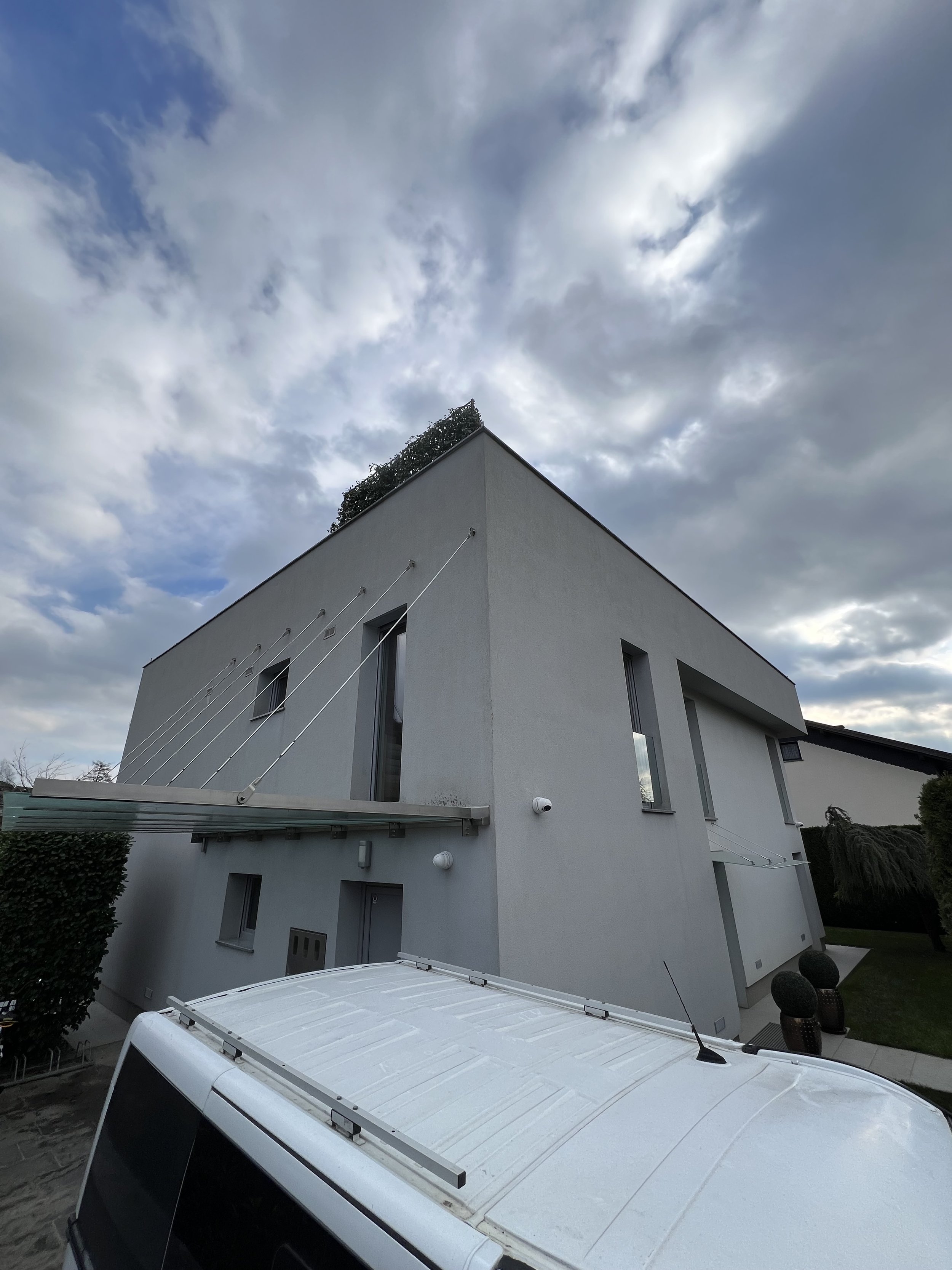 Modern white building with angular design, tall narrow windows, and a rooftop garden, under a cloudy sky, with part of a van visible in the foreground.