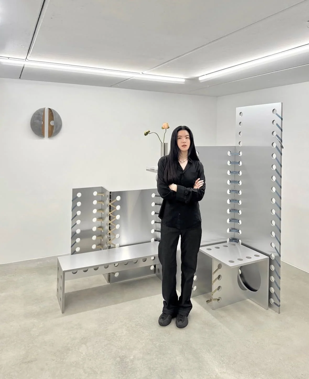 A woman with long black hair in black clothing stands with arms crossed in front of minimalist modern art sculptures in a gallery setting, featuring metallic geometric structures, a white vase with flowers, and a wall clock.