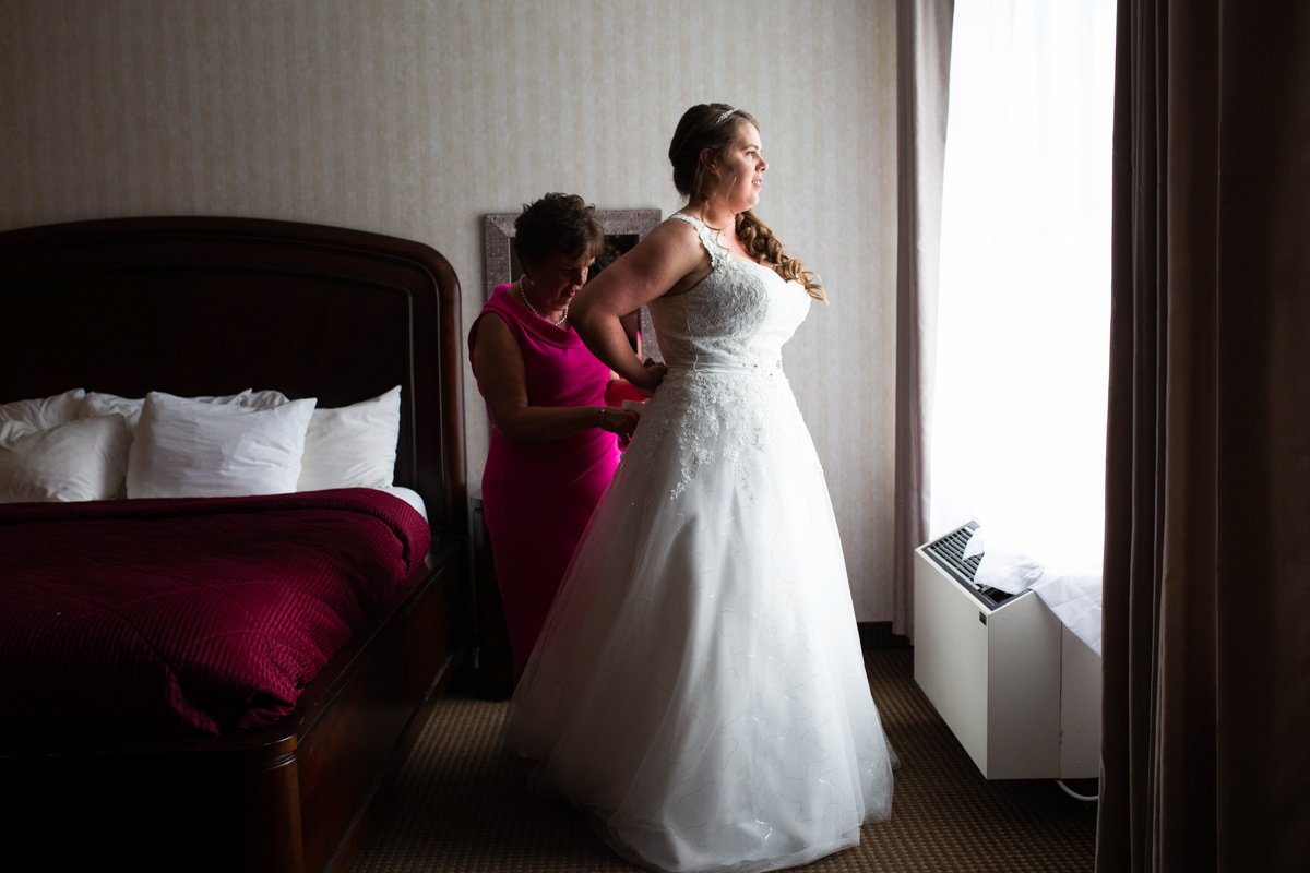 A bride in a white wedding dress standing by a window as her mother helps her with her dress in a bedroom.