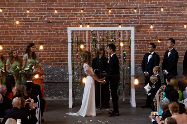 A wedding ceremony with a bride and groom holding hands, standing inside a white frame, with a brick wall backdrop decorated with hanging lights and greenery. Guests are seated on chairs, watching the couple, with some taking photos.