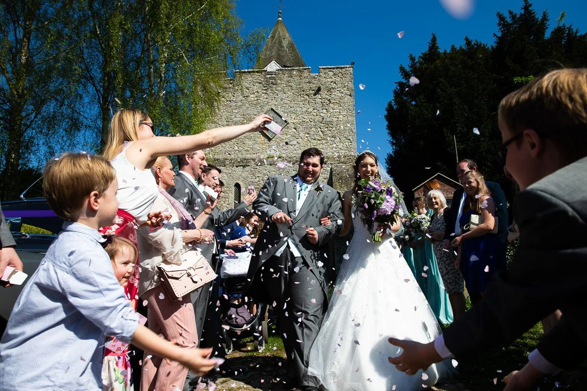 A bride and groom walking through a crowd of guests, tossing flower petals during their wedding celebration outdoors on a sunny day with a castle-like stone building in the background.