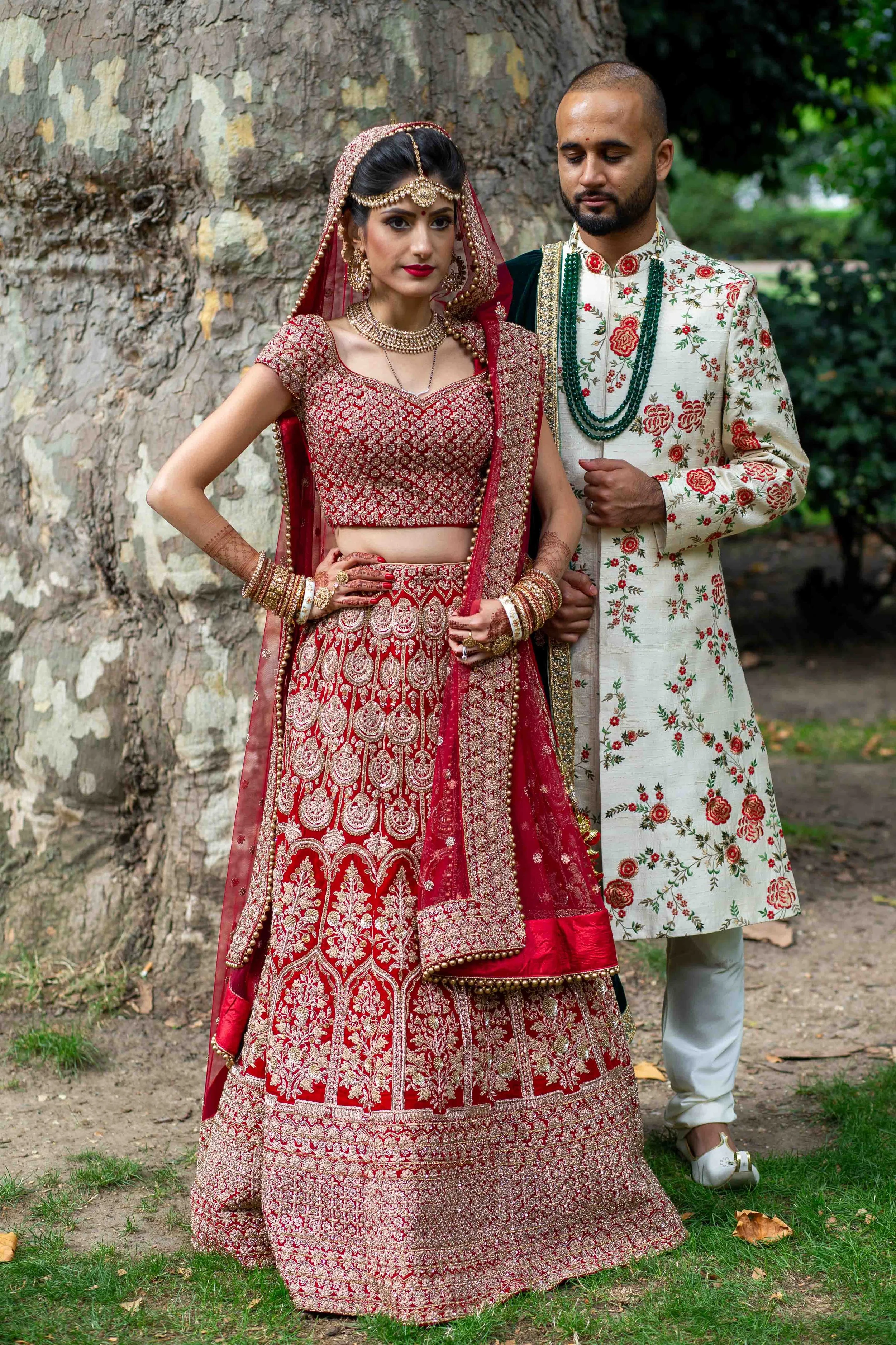 A couple dressed in traditional Indian wedding attire standing outdoors next to a large tree.