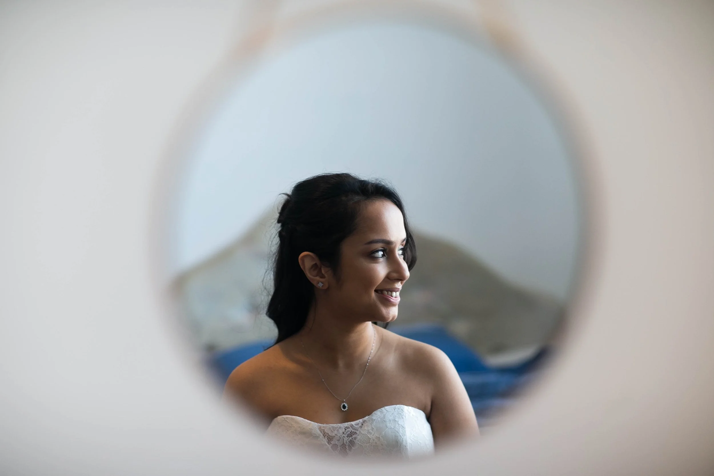A woman with dark hair, wearing a strapless white dress, smiling and looking to her right, seen through a circular reflection or mirror.