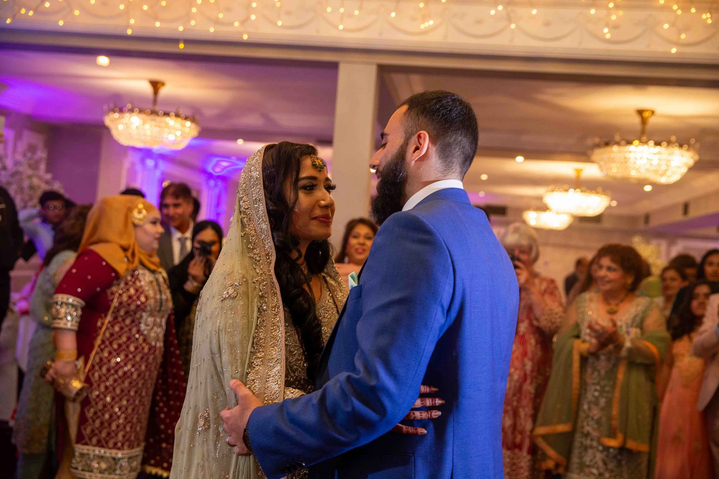 A couple dancing at a wedding reception, surrounded by guests in traditional attire, with ornate chandeliers and soft purple lighting.