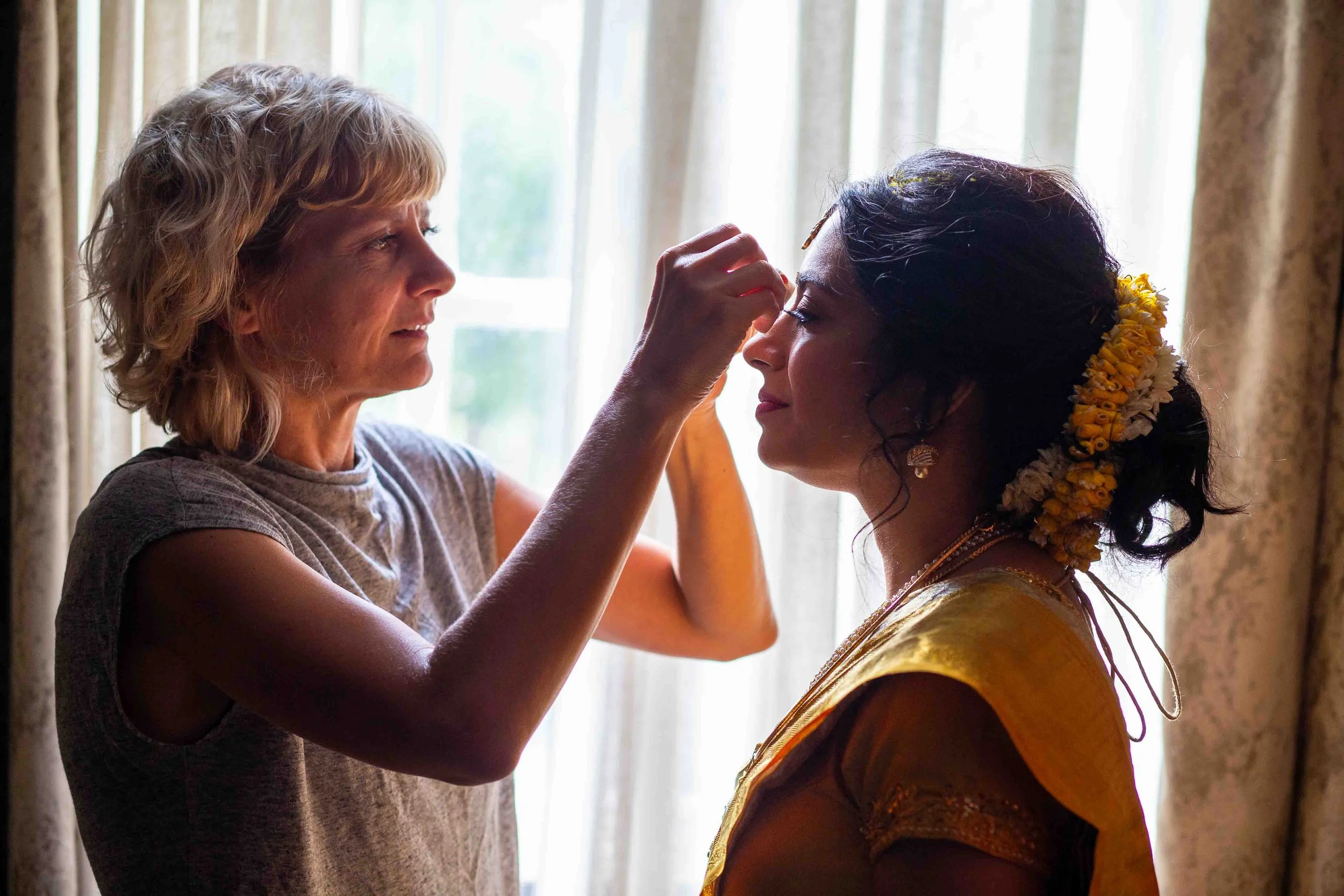A woman with a floral hair accessory is having her face painted or decorated by another woman indoors near a window.