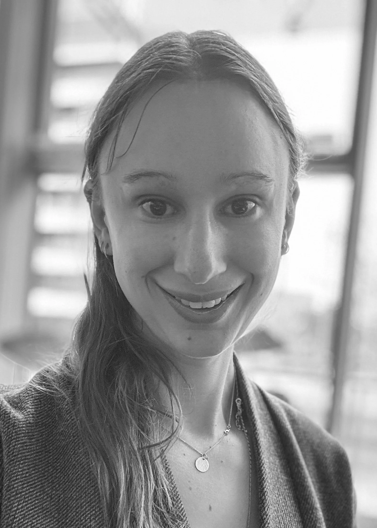 Black and white close-up photo of a smiling woman with long hair, wearing a necklace and earrings, indoors with stairs in the background.