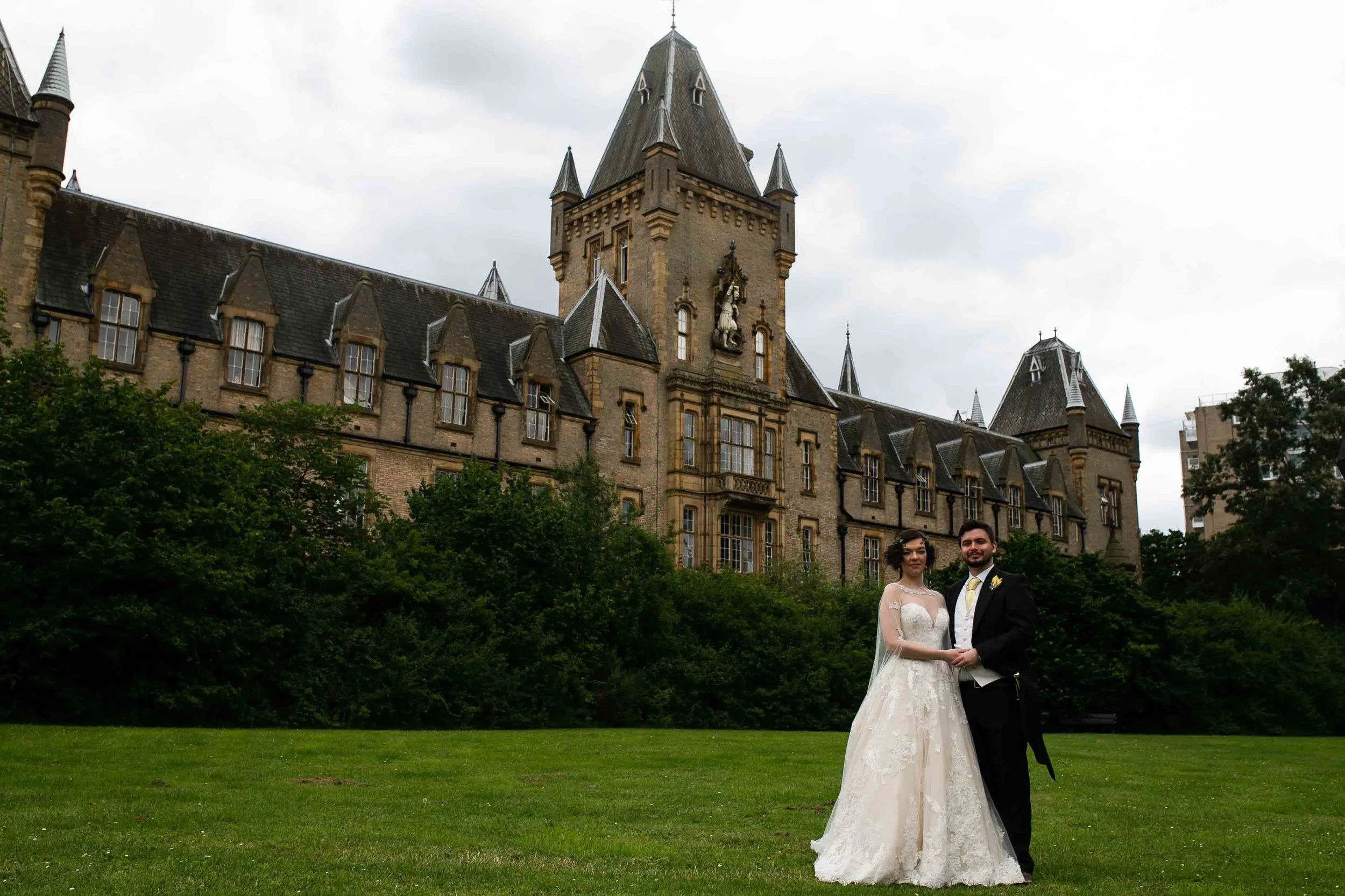 A bride and groom standing on a lawn in front of a historic castle with turrets and a clock tower, greenery in the foreground, and cloudy sky overhead.