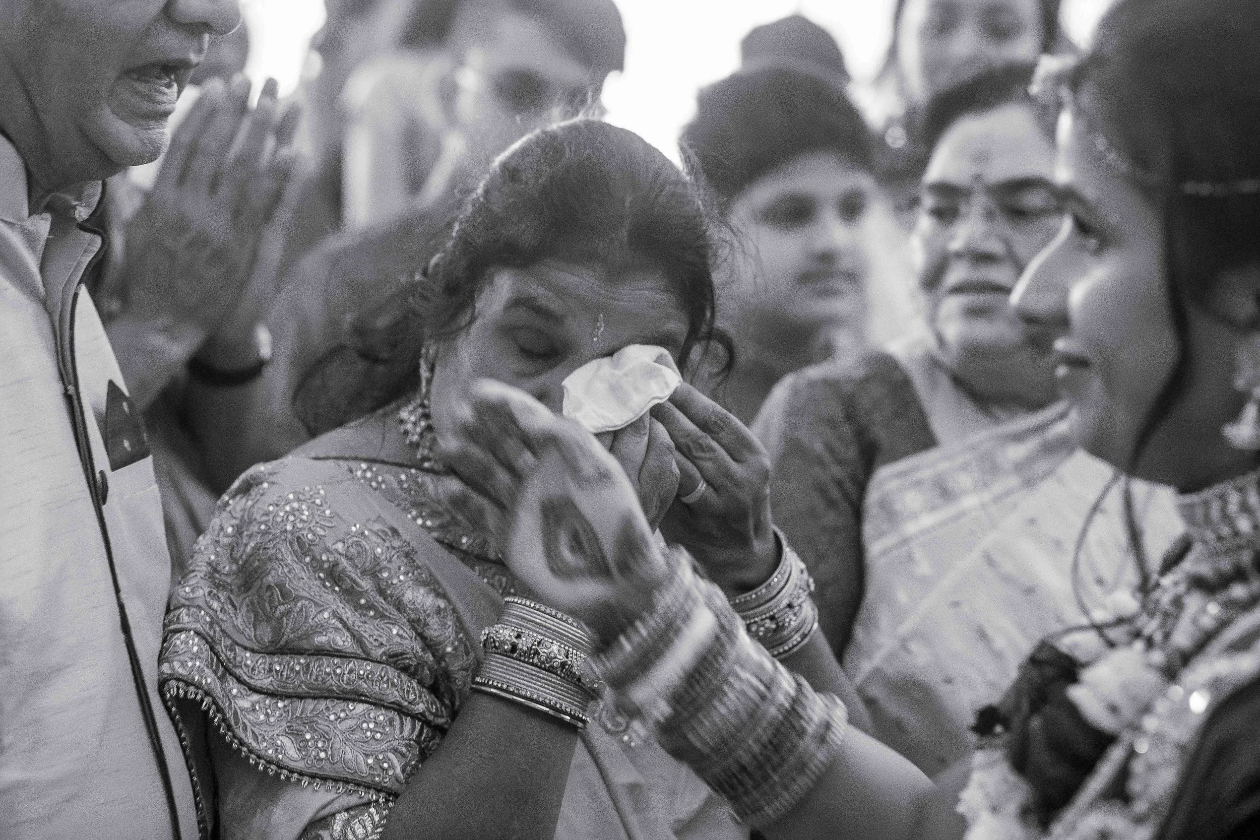 A woman in traditional Indian attire is crying and wiping her tears with a tissue, surrounded by other women and a man, at a ceremonial or emotional event.