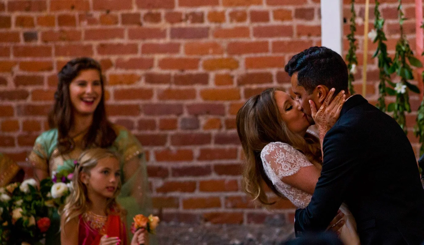A bride and groom kiss at their wedding while seated. Two women, possibly wedding guests, watch in the background, one smiling and holding a bouquet, the other looking on, both dressed in colorful attire. The setting features a brick wall and green f
