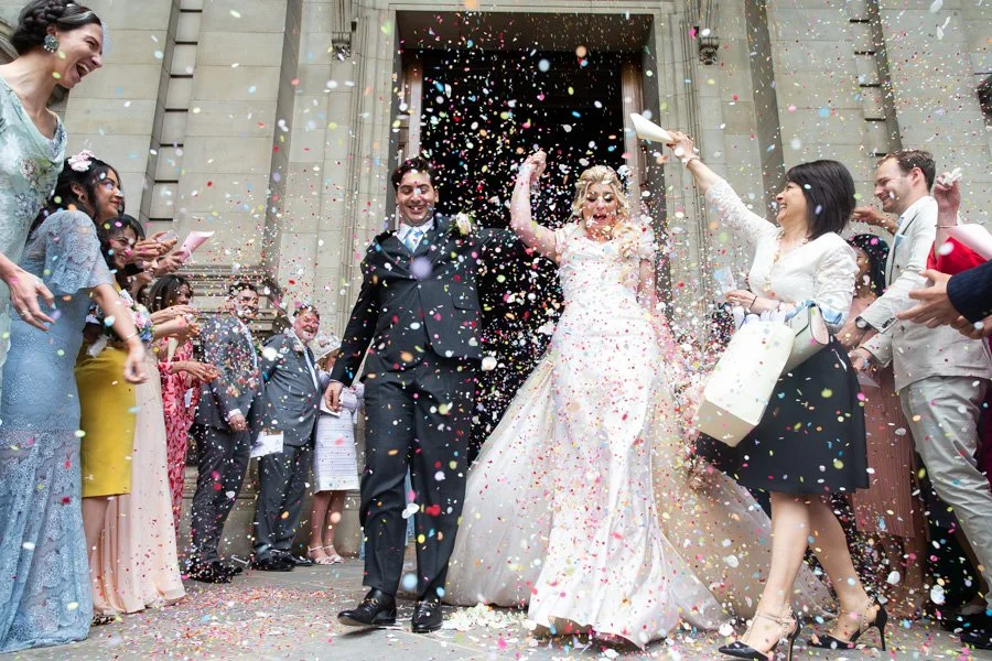 Bride and groom exiting a building while guests throw confetti in celebration.