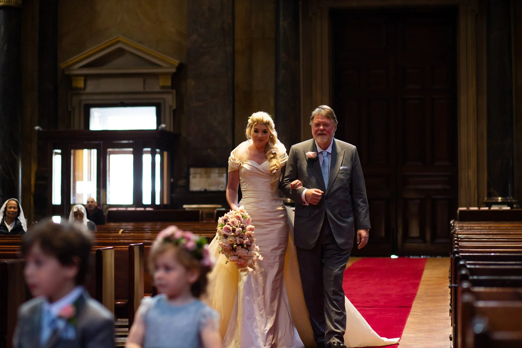 A bride with long blond hair in a cream-colored wedding gown walking down the aisle with an older man in a gray suit and tie, inside a church with wooden pews and a red carpet.