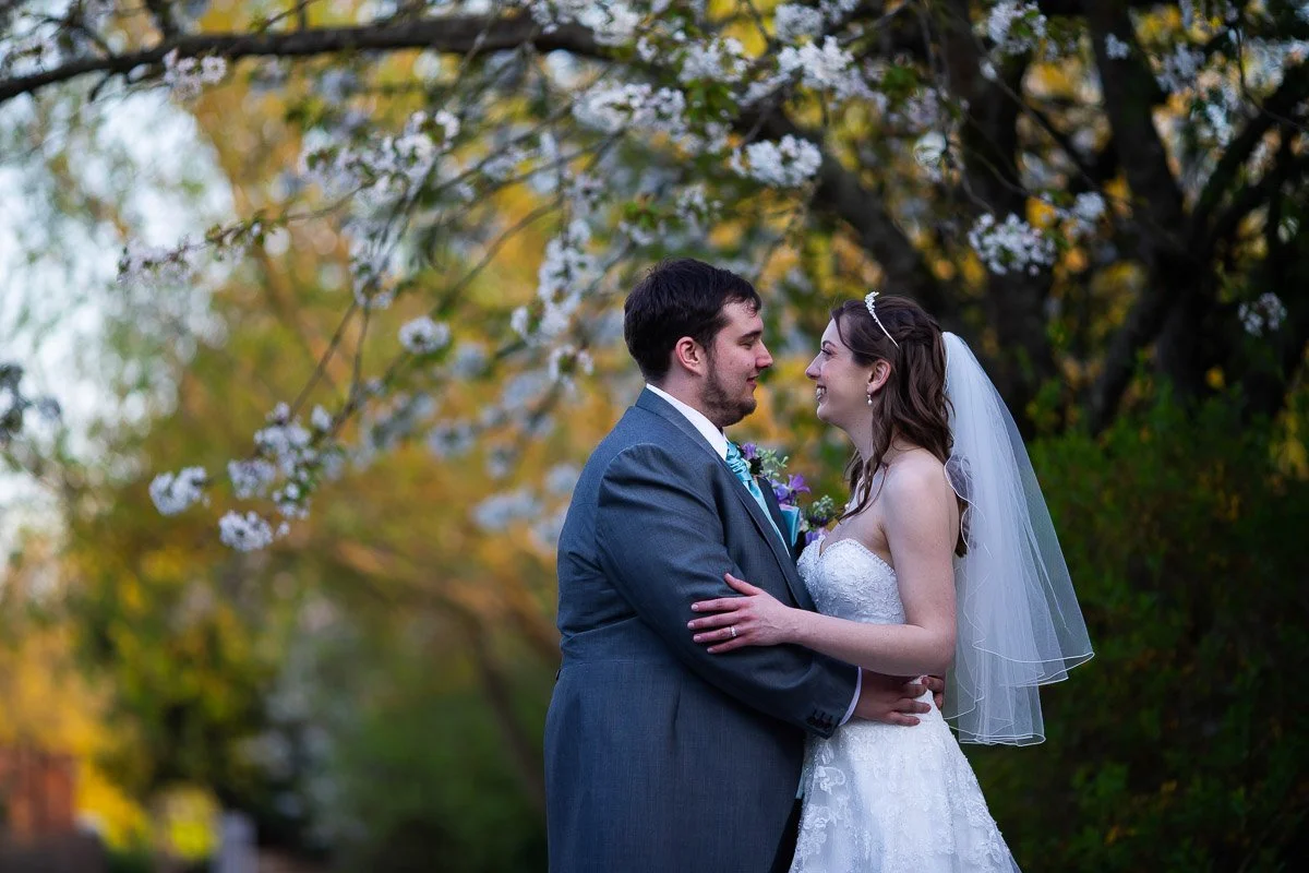 A newlywed couple stands close under blooming trees, gazing into each other's eyes on their wedding day.