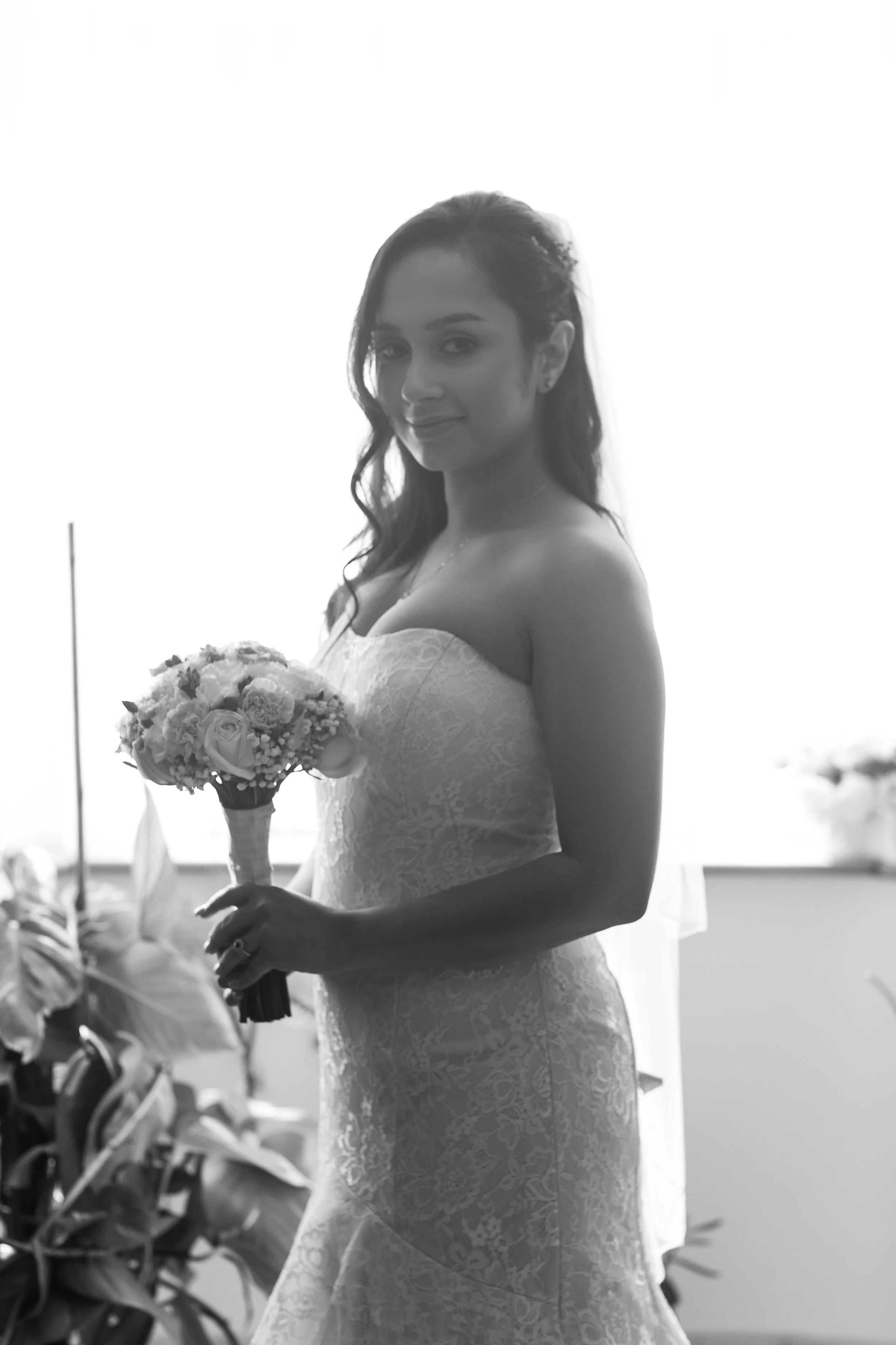 A woman in a lace wedding dress holding a bouquet of flowers, smiling softly, with plants and a blurred outdoor background.