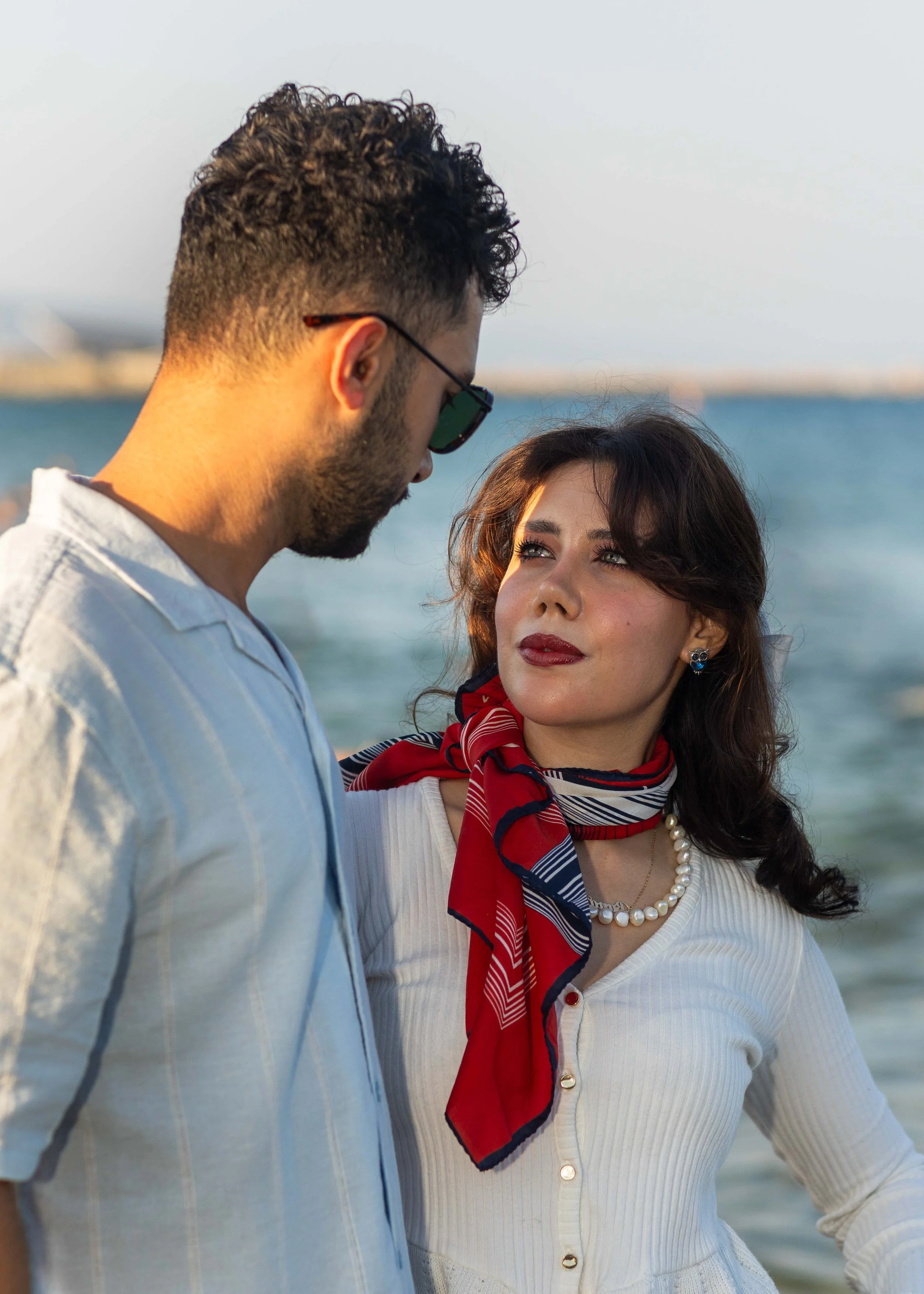 A man and a woman are standing close together outdoors by the water, with the man looking down at the woman and the woman looking up at the man.