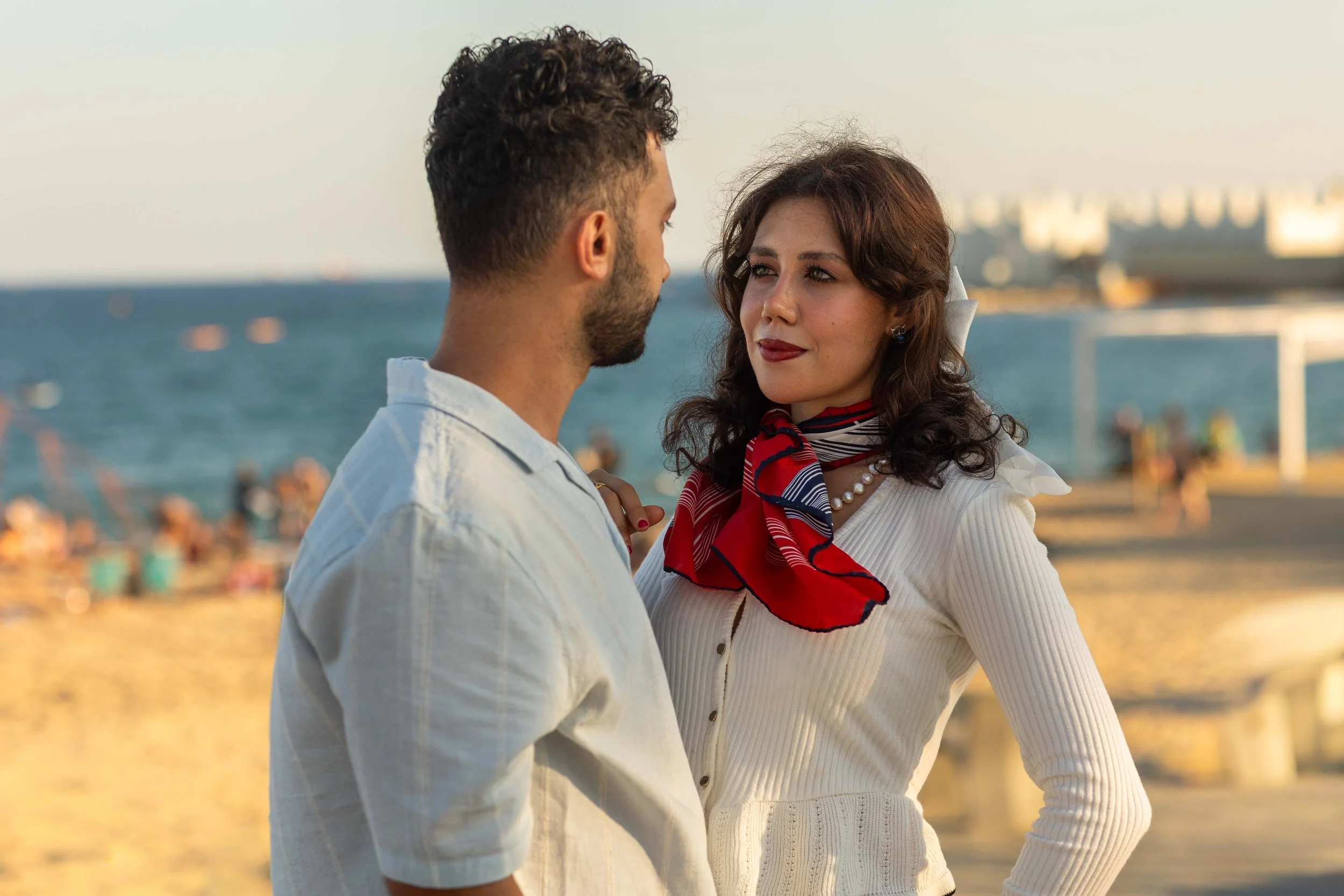 A man and woman facing each other on a beach, with water and blurred city buildings in the background.