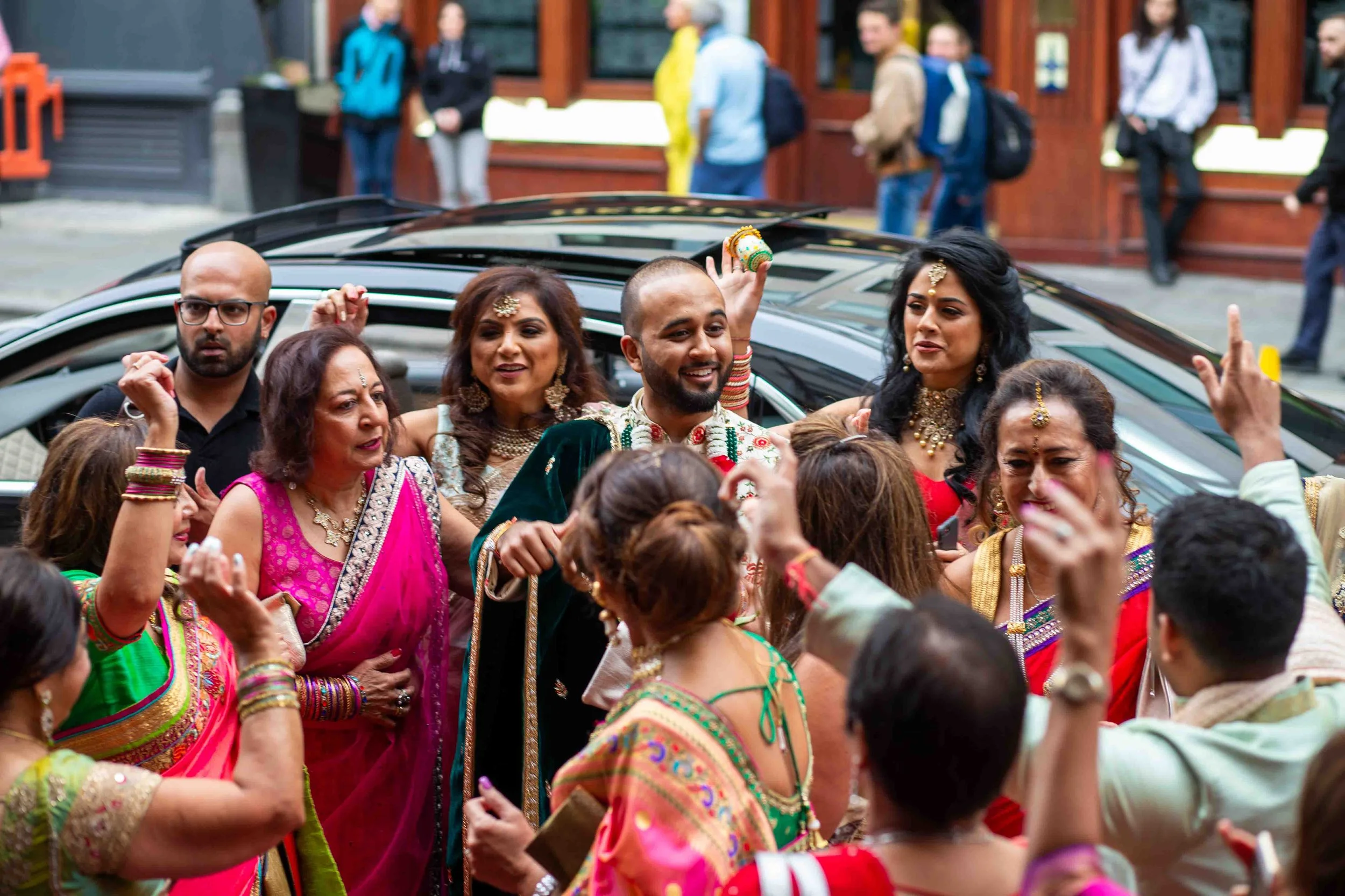 A group of people dressed in colorful traditional Indian attire dancing and celebrating outdoors, with a man in the center smiling and holding a ring, surrounded by women in sarees with jewelry.
