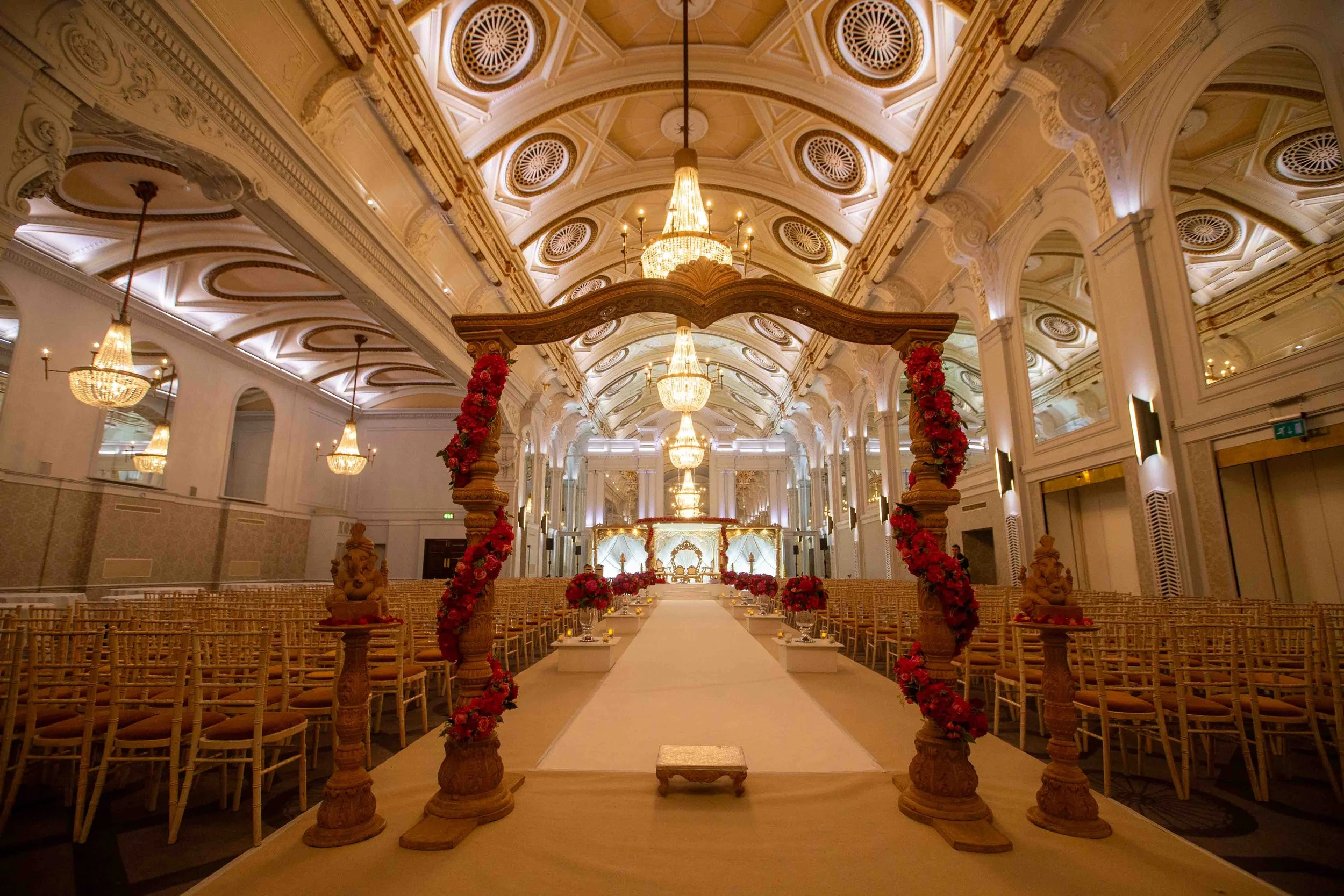 Elegant indoor wedding ceremony setup with a white aisle, decorated with red flowers and statues, leading to an ornate altar with chandeliers and gold accents in a grand hall.